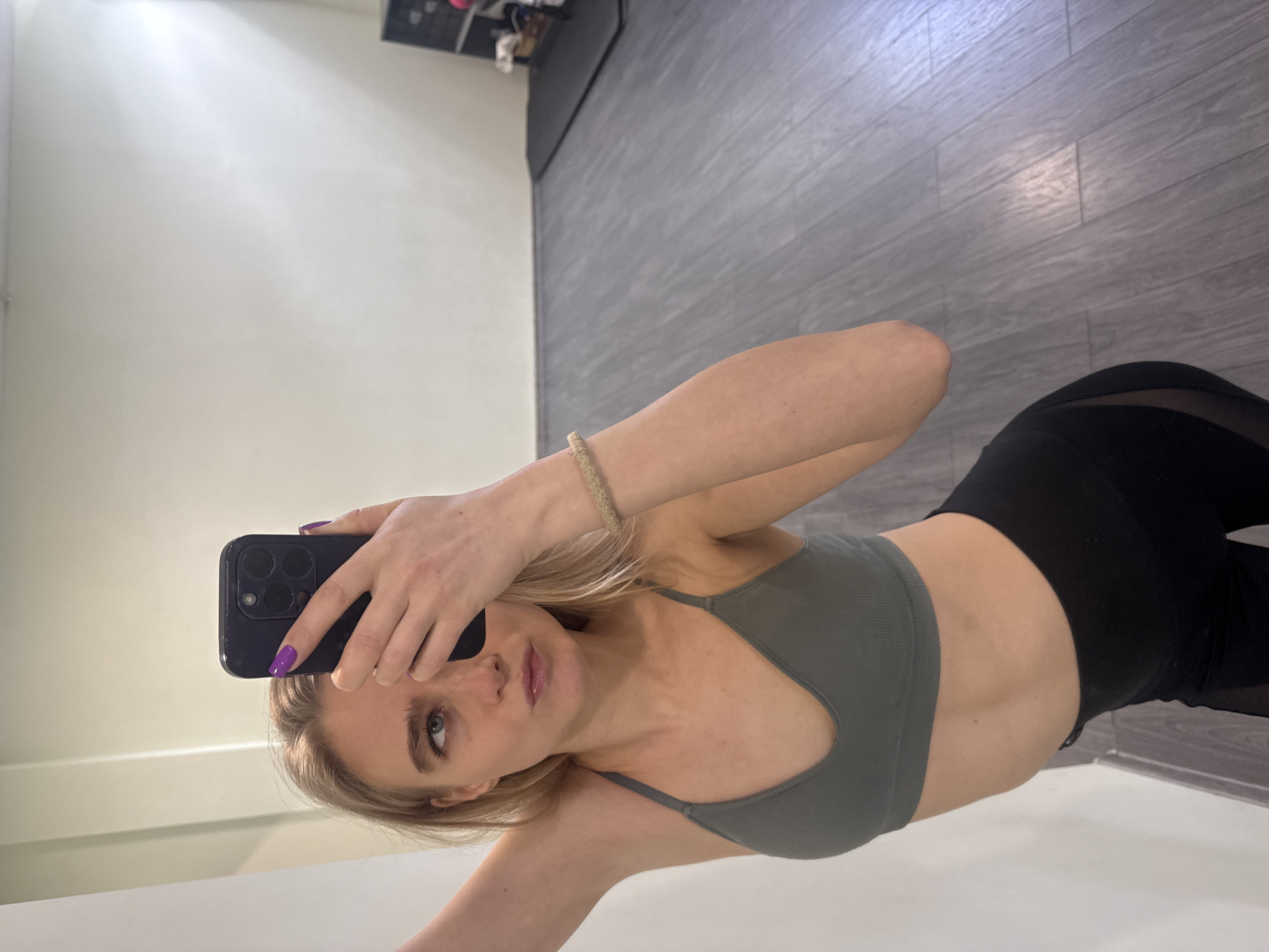 A young woman is captured in a mirror selfie, holding a dark-colored smartphone to take the picture. She is positioned centrally in the frame, looking directly into the reflection. Her left hand, adorned with bright purple nail polish, holds the phone, partially obscuring the upper part of her face.
The woman has blonde hair, light-colored eyes, and a neutral expression with slightly parted lips. She is wearing a ribbed, olive-green sports bra or tank top that exposes her midriff, paired with black leggings. A light-colored, possibly braided, bracelet is visible on her right wrist.
The background consists of a plain, light-colored wall on the left and a floor with grey, wood-like planks on the right. In the upper right corner of the background, some indistinct dark objects or furniture are visible. The lighting is bright and even, suggesting an indoor setting like a gym, studio, or private room. There is no visible text in the image.