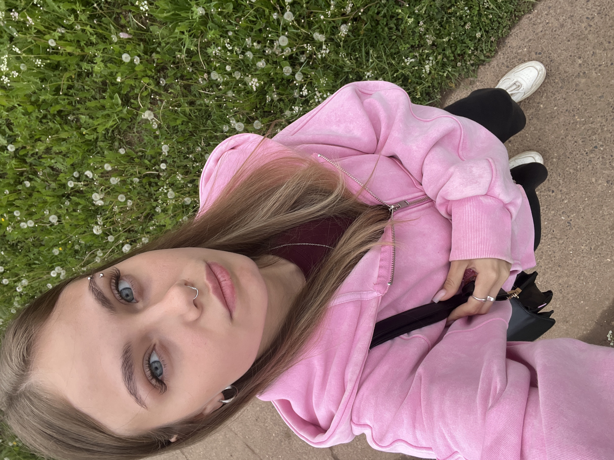A young female is depicted outdoors, looking directly upward into the camera. She has long, light brown hair, blue eyes, and fair skin. Her right eyebrow features a triple-ball silver curved barbell piercing, her nasal septum a wavy silver ring, and her left ear a small silver hoop earring. She is wearing a faded pink full-zip hoodie, with its hood up but not covering her head, over a dark red long-sleeved shirt, black pants, and white low-top sneakers. Her right hand, adorned with a silver square ring on the index finger and light pink manicured nails, holds a small black shoulder bag with a gold clasp. She is positioned on a light grey concrete or asphalt path. Filling the right side and background of the image is a patch of green grass densely populated with white dandelion seed heads and smaller white flowers. The scene is illuminated by natural daylight. This image was captured in the city of Фоки, Russia.