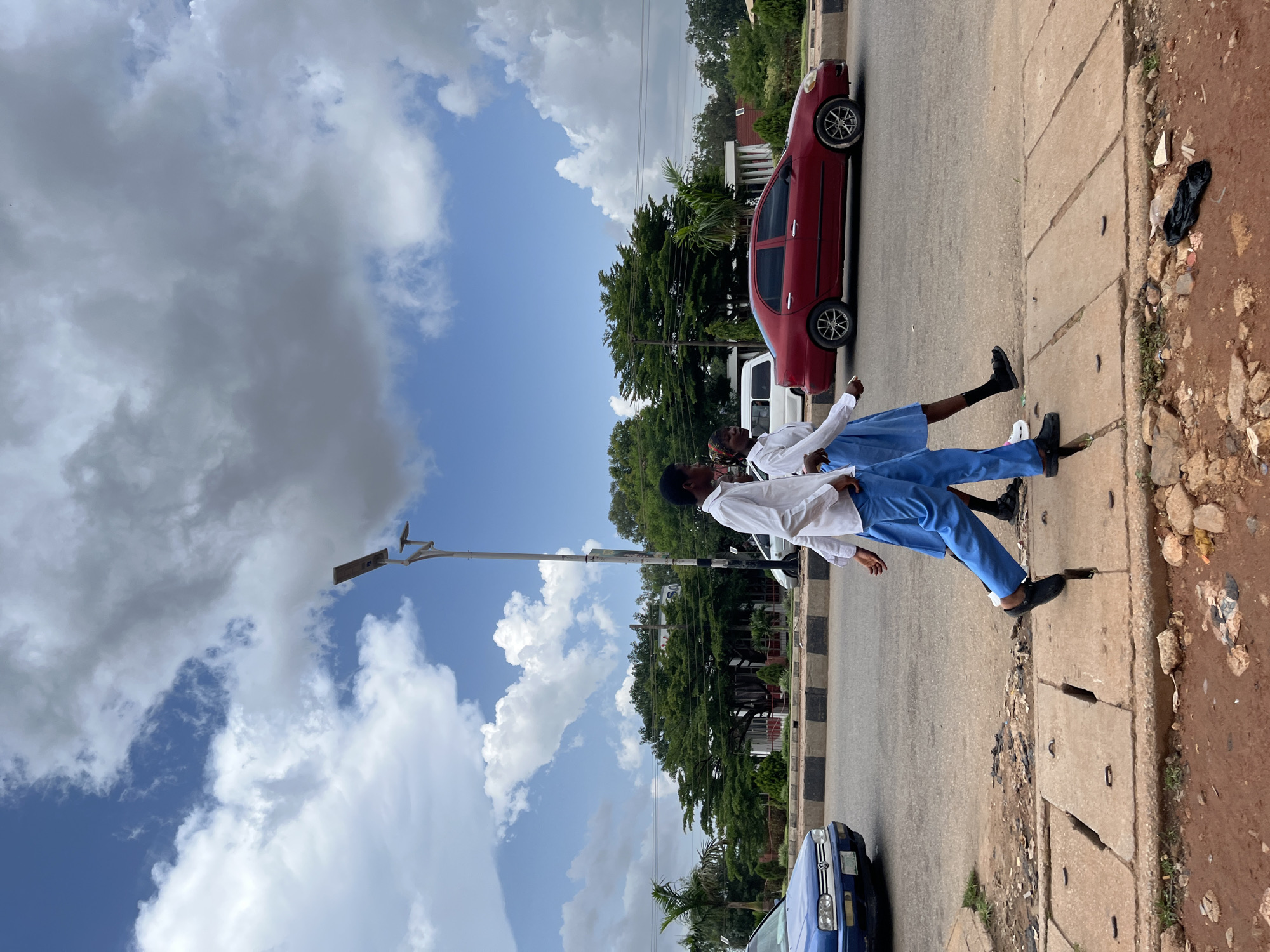 Two young individuals, likely students in uniform, are walking on a sidewalk next to a road. The individual in the foreground is wearing a white shirt and blue pants, with black shoes and socks. Their hair is styled in braids. The second individual, slightly behind and to the left, is also in a white shirt and blue pants, with black shoes. They appear to be looking away from the camera.
A red car is parked on the side of the road, and a white vehicle is visible behind it. A blue car is also parked on the road in the foreground. The scene is set outdoors, with a sidewalk made of concrete slabs and a dirt and gravel area to the right, which shows some litter. Trees and buildings are visible in the background, suggesting an urban or semi-urban environment. A street light pole with a solar panel is also present.
The sky is predominantly blue with scattered white and grey clouds, indicating it is daytime, possibly late morning or early afternoon. The weather appears to be bright, with the sun casting shadows. The overall mood seems casual, with the subjects appearing relaxed as they walk. There is no visible text in the image. The context provided places the scene in Jos, Nigeria.
