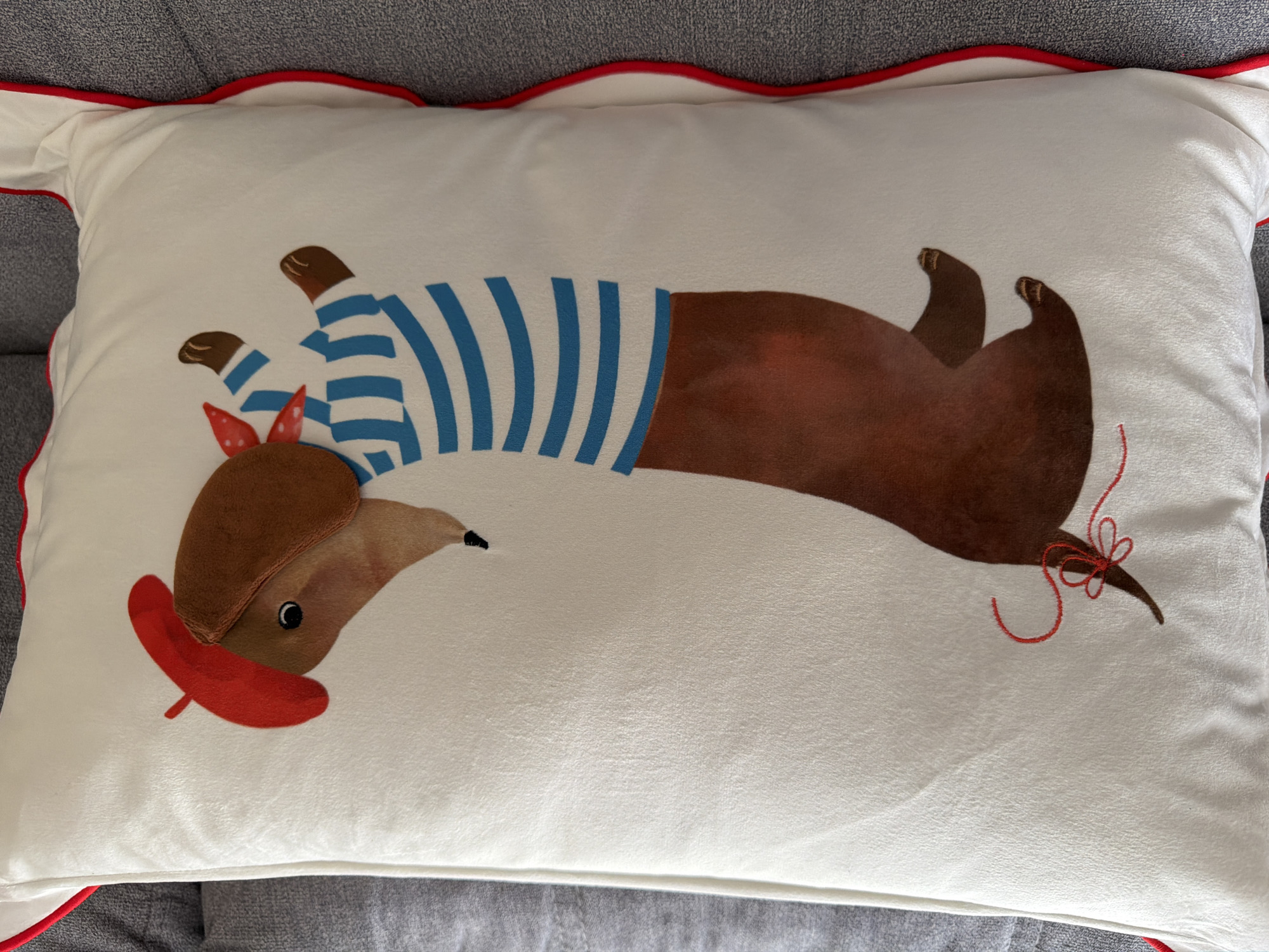A white throw pillow with a detailed illustration of a dachshund dog is displayed on a gray sofa. The dog is depicted in profile, facing right, wearing a red beret on its head and a blue and white striped shirt. A red bandana is tied around its neck. The dog's tail is curled with a red string bow. The pillow has a scalloped red trim along its edges. The image is an indoor shot, likely a living room or bedroom, with the pillow serving as a decorative item. The location in Pazardzhik, Bulgaria, is not specified or inferable from the visual information provided.