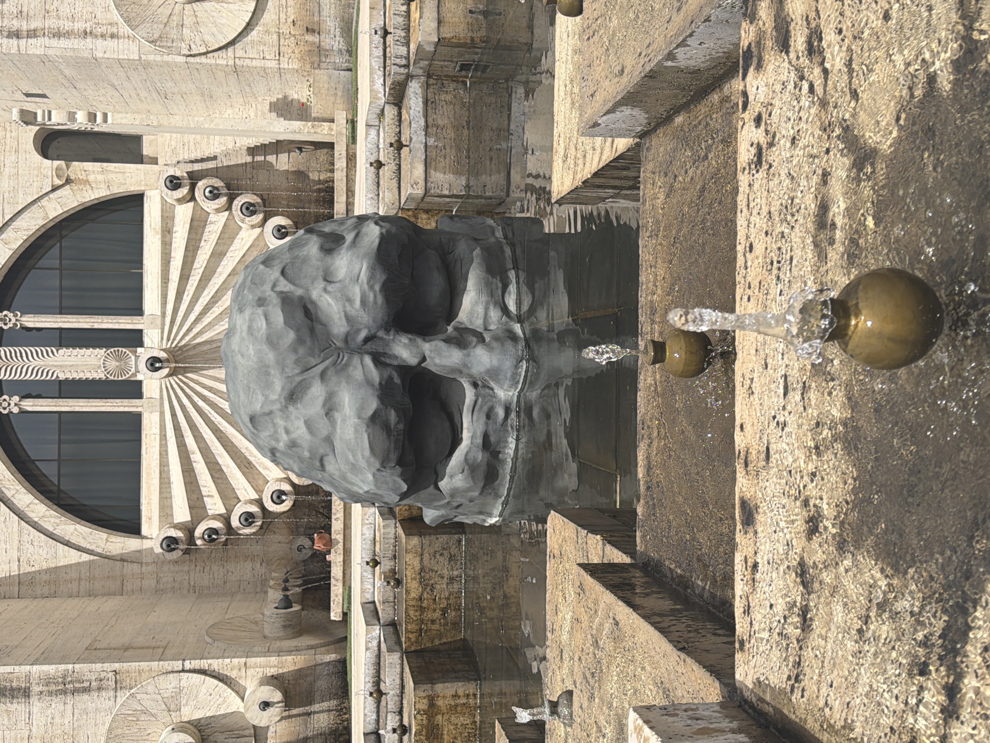 The image shows a view of the Cascade in Yerevan, Armenia. The Cascade is a monument with stairs, fountains, and sculptures. In the foreground, a bronze orb releases water. Beyond that, a gray rock sculpture in the shape of a face sits in the water. The architectural features of the Cascade with its series of steps and decorative elements are in the background. A person is visible standing near the sculpture. The lighting suggests it is daytime with some shadows present.
