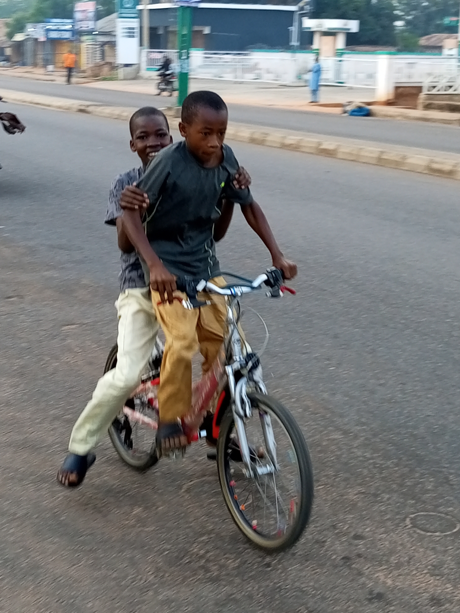 Two young boys are riding a bicycle on a paved road, likely in the city of Gombe, Nigeria. The boy in the front is wearing a dark grey t-shirt and khaki pants and is operating the bicycle. The boy in the back, wearing a grey patterned t-shirt and light-colored pants, is sitting behind the front boy, with his arms around him. They are both wearing sandals. The bicycle has a red frame and silver handlebars. The road surface appears to be asphalt, with a concrete curb on the right side. In the background, there are buildings with signs and some trees. The lighting suggests it is either early morning or late afternoon.