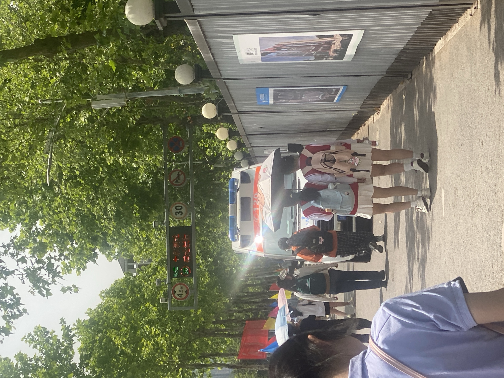 The image depicts a street scene in what appears to be the Chongqing High-tech Zone in China. The street is lined with trees on the left, and a corrugated metal fence runs along the right side, with posters displayed on it. An ambulance is parked on the street with people standing around it. Pedestrians are walking on the sidewalk. A street sign indicating a speed limit of 30 is visible, as is an electronic sign displaying the current time and temperature. Colored flags are strung along the street.