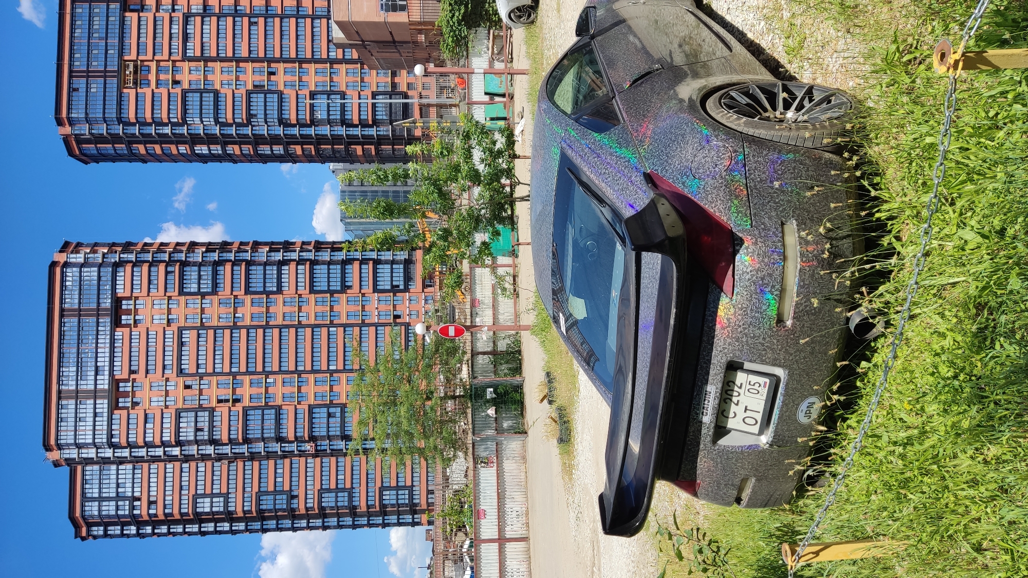 In Krasnodar, Russia, a holographic-wrapped sports car, possibly a Nissan 350Z given its distinct rear spoiler and taillight design, is parked on a gravel lot overgrown with grass. The car features a large rear wing and a license plate reading "C 202 OT 05". In the background, two identical, modern high-rise residential buildings with orange brick facades and dark-framed windows dominate the skyline under a bright blue sky with scattered clouds. A construction fence with warning signs and a "No Entry" sign with a red circle and a horizontal bar is visible between the car and the buildings. A small portion of another white car is visible parked to the right of the sports car. The scene depicts an urban environment undergoing development, with a focus on the unusual car and the imposing architecture.