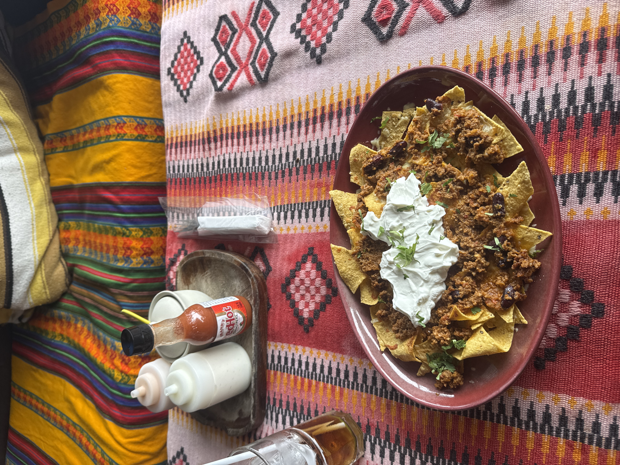 The image depicts a plate of nachos with ground meat, black beans, sour cream, and cilantro on a red plate placed on a colorful patterned tablecloth. Next to the plate, there is a wooden tray containing condiments, including a bottle of Frank's Red Hot sauce, two squeeze bottles with white sauce, and a small white container with a yellow straw. There's also a package of white napkins, a glass with a straw, and colorful pillow(s) on the left side of the image. The scene is photographed indoors. The location is Thong Sala, Thailand.