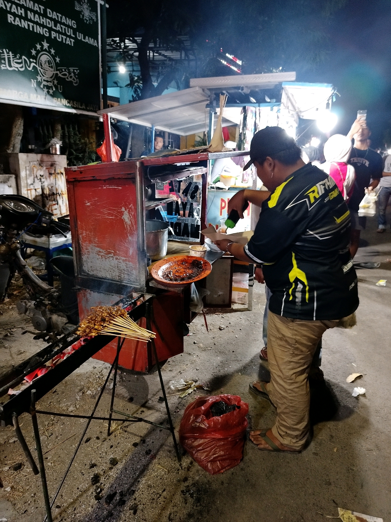 An outdoor food stall operates at night on a street in Putat, Indonesia. A man wearing a black cap and a black and yellow patterned shirt is cooking satay over a charcoal grill. Satay skewers are visible on the grill and on a metal rack. A red cabinet is part of the food stall. Other people are in the background, some holding up phones. The ground is paved and littered with debris. A red plastic bag sits on the ground. A green banner with Arabic writing is visible in the upper left corner of the image.