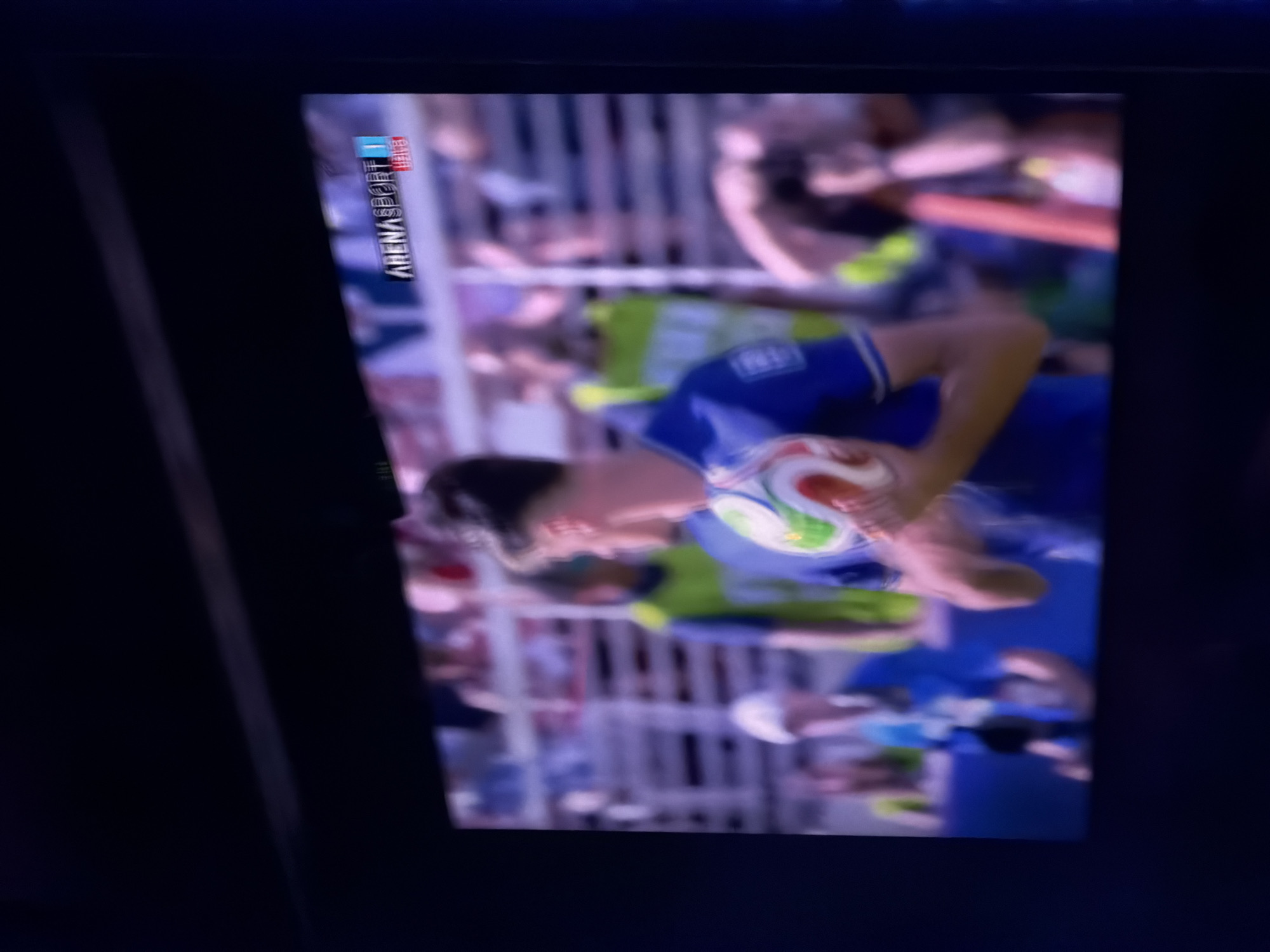 The image shows a close-up of a television screen displaying a live sports broadcast. The broadcast appears to be a soccer match, with a player in a blue jersey holding a soccer ball. The logo for "ARENASPORT 1" is visible in the top right corner of the screen. The background of the broadcast shows blurred images of spectators in a stadium. The television screen itself is framed by a dark border, suggesting it is set within a dimly lit room. The overall impression is that of someone watching a game on television. The prompt's request to include the location of Travnik, Bosnia and Herzegovina, cannot be fulfilled as there is no visual information in the image to suggest this specific location.