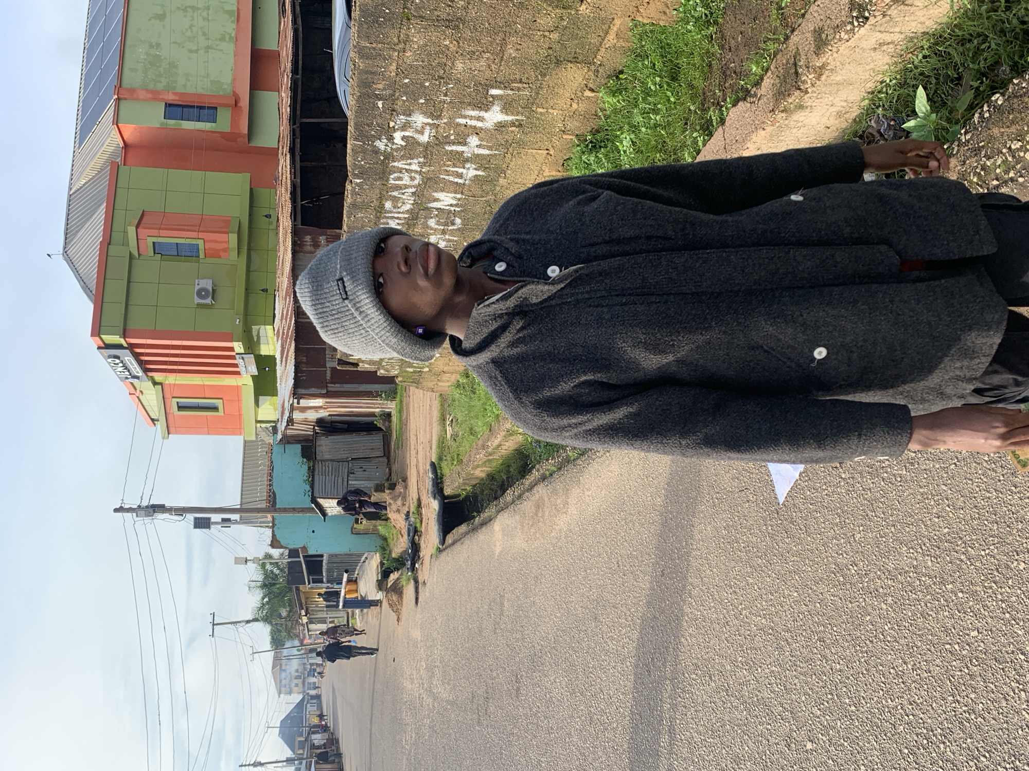 The image depicts a street scene in Jos, Nigeria. In the foreground, a young Black male stands facing right, looking away from the camera. He is wearing a grey knit beanie and a dark grey zip-up jacket over a red garment. His right hand is partially visible, holding a small object. Behind him, a concrete wall is visible with white lettering that appears to read "WIGABA 2" and "ACM 4++".
The background shows a paved street with a drainage ditch running along the right side. Several people are walking or standing further down the street. Buildings line both sides of the street, including a multi-story structure with orange and green facade. Power lines and poles are prominent in the sky. The overall environment suggests a somewhat busy urban setting with elements of commercial and residential buildings. The lighting indicates it is daytime, possibly late morning or early afternoon, with clear sky and shadows cast.