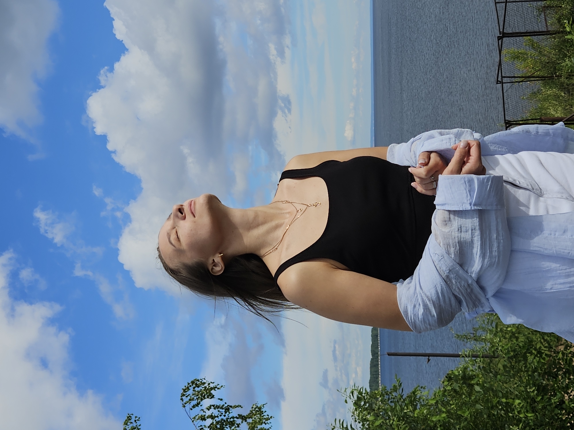 A young woman stands with her eyes closed, head tilted back as if enjoying the sun. She wears a black tank top, white pants, and a light blue open shirt draped over her arms. Her hair is light brown and windswept. She is positioned in the foreground, with a wide body of water and a tree line in the background. The sky is blue with scattered white clouds. The scene appears to be outdoors, possibly a park or viewpoint overlooking the Kama River, in the city of Chaikovsky, Russia.