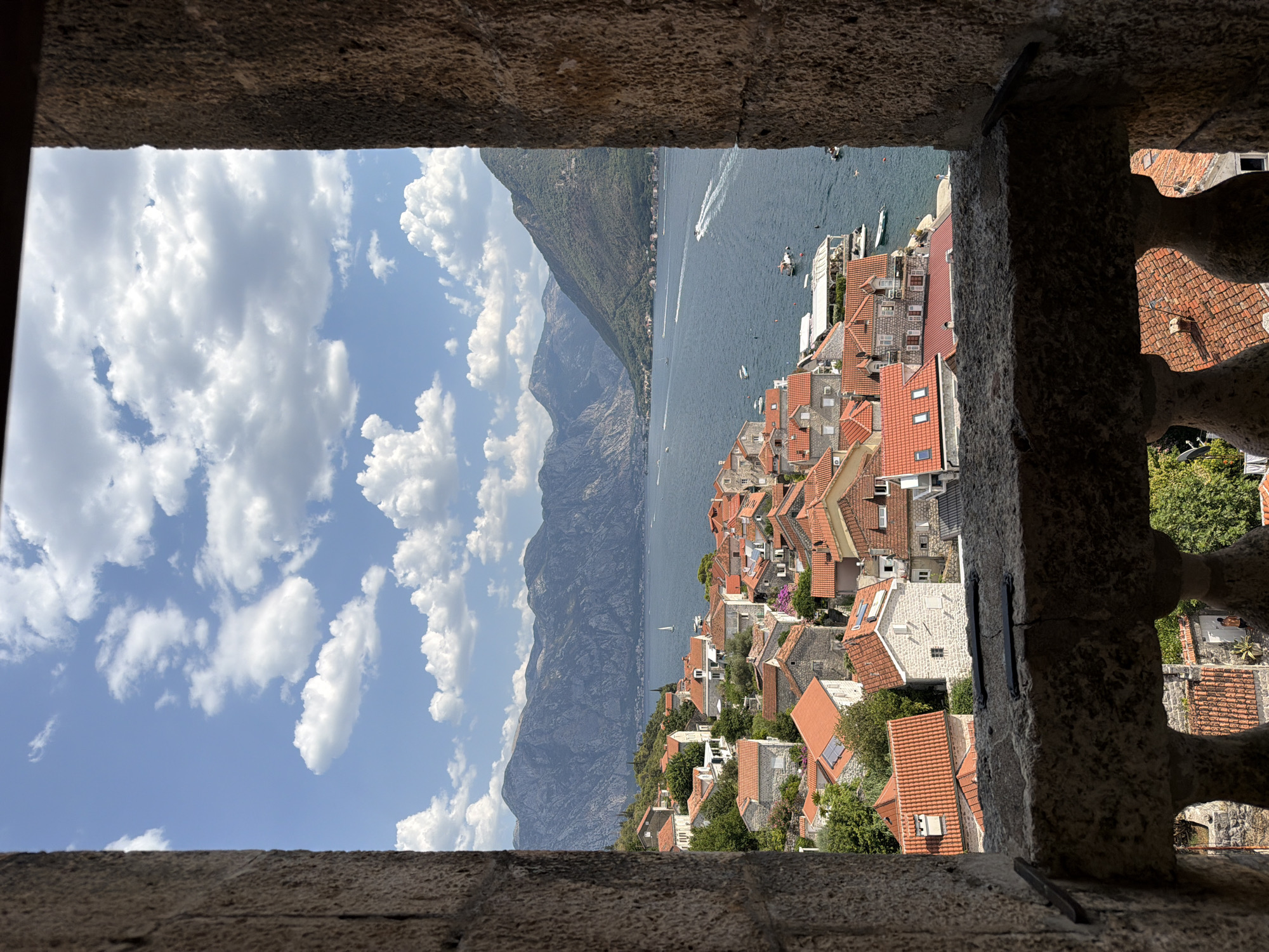 This image depicts a picturesque view of the town of Perast, Montenegro, as seen through a stone window frame. The foreground is dominated by the rough, textured stone of the window structure, which frames the scene. A stone balcony with decorative balusters is visible below the window opening.
Beyond the frame, the town of Perast unfolds with its characteristic terracotta-tiled roofs cascading down the hillside towards the Bay of Kotor. Numerous buildings with red and orange roofs are densely packed together, interspersed with patches of green foliage. A prominent white building with a light-colored roof stands out among the others.
The deep blue water of the bay stretches out, dotted with several boats. Some are small motorboats, leaving white wakes on the water, while others appear to be moored or moving at a slower pace. A single white sailboat is visible in the middle distance.
In the background, imposing mountains rise, their slopes covered in lush greenery, creating a dramatic backdrop to the coastal town. The sky above is a vibrant blue, adorned with fluffy white cumulus clouds. The overall scene conveys a sense of tranquility and historical charm, characteristic of the Mediterranean coastal towns.