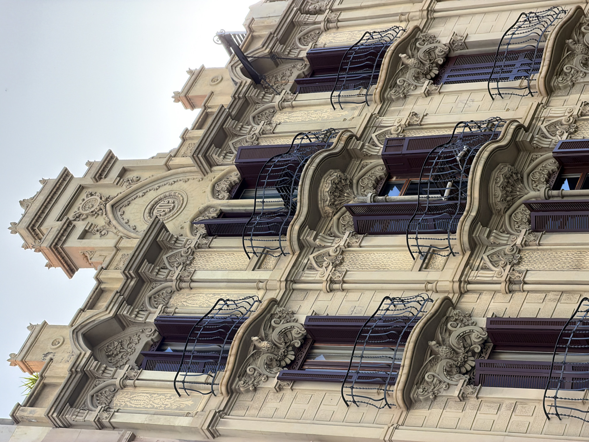 This is a close-up, low-angle exterior, eye-level shot of the highly ornate facade of a historic building, likely residential, in Barcelona, Spain. The building is made of light-colored stone and features elaborate architectural details. Several floors are visible, each adorned with balconies that have intricate wrought-iron railings. The balconies are supported by decorative corbels, one of which features a sculpted bird-like creature with outstretched wings.
The windows on each floor are covered with dark brown louvered shutters, some of which are partially open, revealing glimpses of glass. The facade itself is decorated with bas-relief carvings of floral motifs, scrollwork, and geometric patterns. Above the windows, there are more elaborate decorative elements, including pediments and niches. A circular medallion with the number "1909" is visible on the upper part of the facade, indicating the year of construction or renovation. The sky above is a pale, overcast gray. The architecture suggests a style such as Art Nouveau or Modernisme, common in Barcelona.
