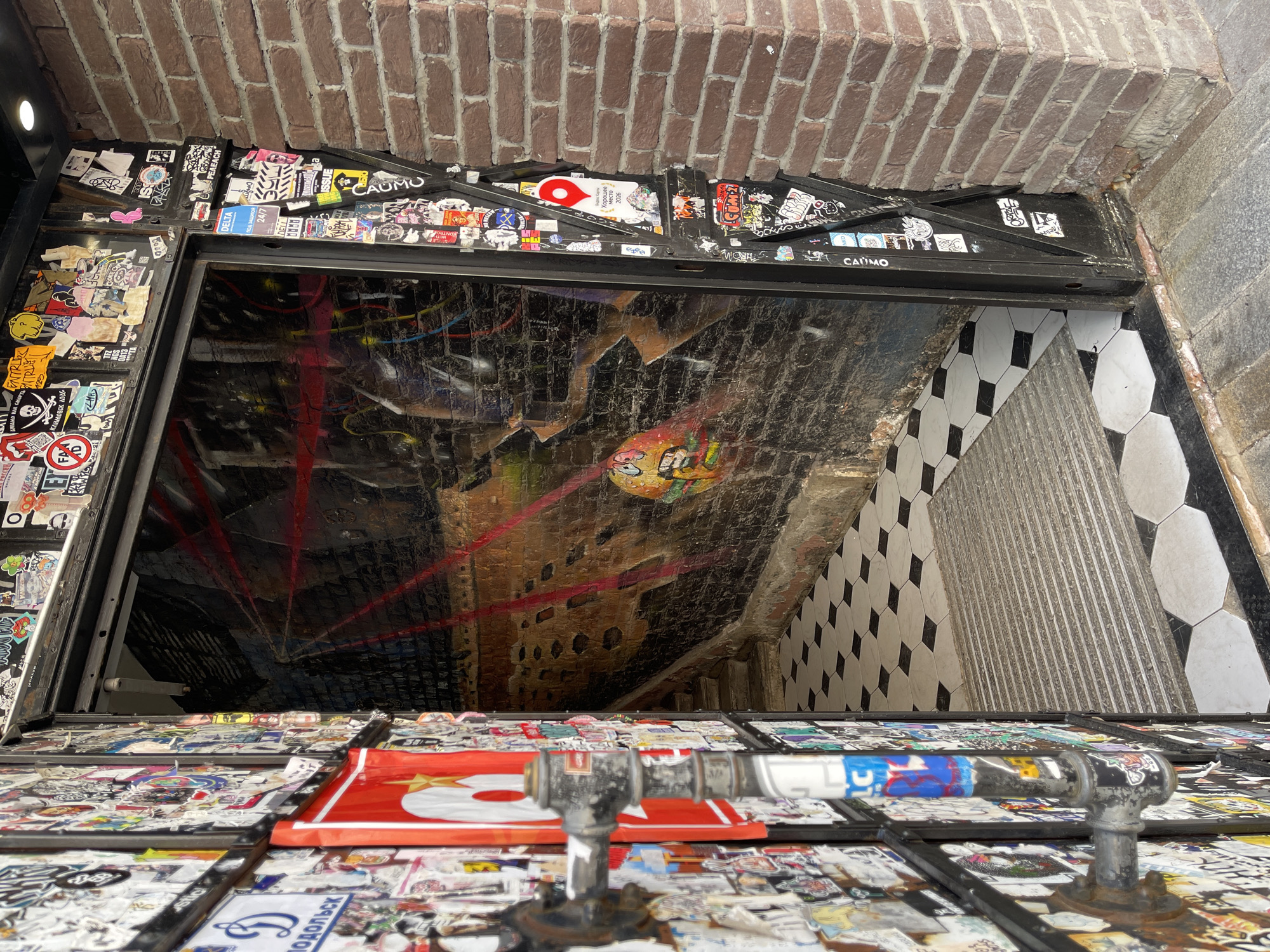 This image shows a view down into an entrance covered with stickers, likely located in Moscow, Russia. The entrance is an opening in a brick structure, covered in numerous stickers on the surrounding framework. Below the entrance is a view of a tiled staircase and interior walls. Graffiti art is visible on the walls, including an image of a cartoonish face. A metal pipe fixture with stickers on it is in the foreground. The environment looks urban and possibly underground. Signs in Russian are present.