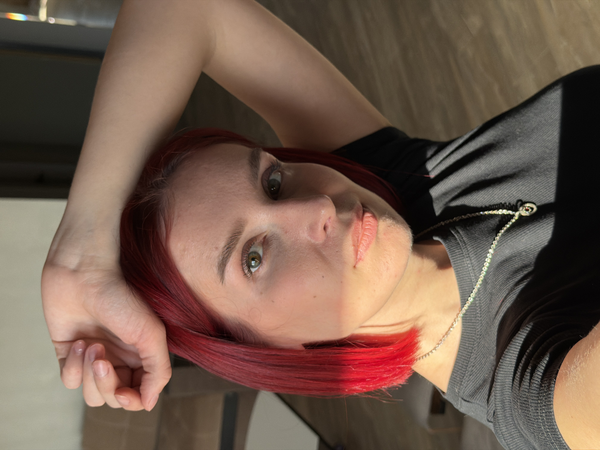 A close-up portrait features a young person, likely female, with striking, vibrant red hair, gazing directly at the camera with a calm and neutral expression. The image is oriented such that the person's head is on the left side of the frame, with their body extending towards the right. One arm is bent, with the hand resting near the top of their head.
The person has fair skin, visible freckles or sun spots, and greenish-brown eyes. They are wearing a black ribbed top or t-shirt, accessorized with a thin silver chain necklace that has a small, indistinct charm.
The scene is characterized by strong, direct sunlight, casting dramatic, sharp shadows across the person's face, neck, and clothing, creating high contrast. Parts of the face, such as the nose, cheekbone, and lips, are brightly illuminated, while other areas are in deep shadow. Similar shadow patterns are visible on the background, which appears to be an indoor setting with a light-colored floor (possibly wood or tile) and darker areas that could be furniture or walls. The lighting suggests a sunny time of day, possibly morning or late afternoon, with light entering through a window. A slight rainbow refraction is visible in the upper left background, hinting at light passing through glass.
No discernible text is visible in the image. The setting is in Chaikovsky, Russia.