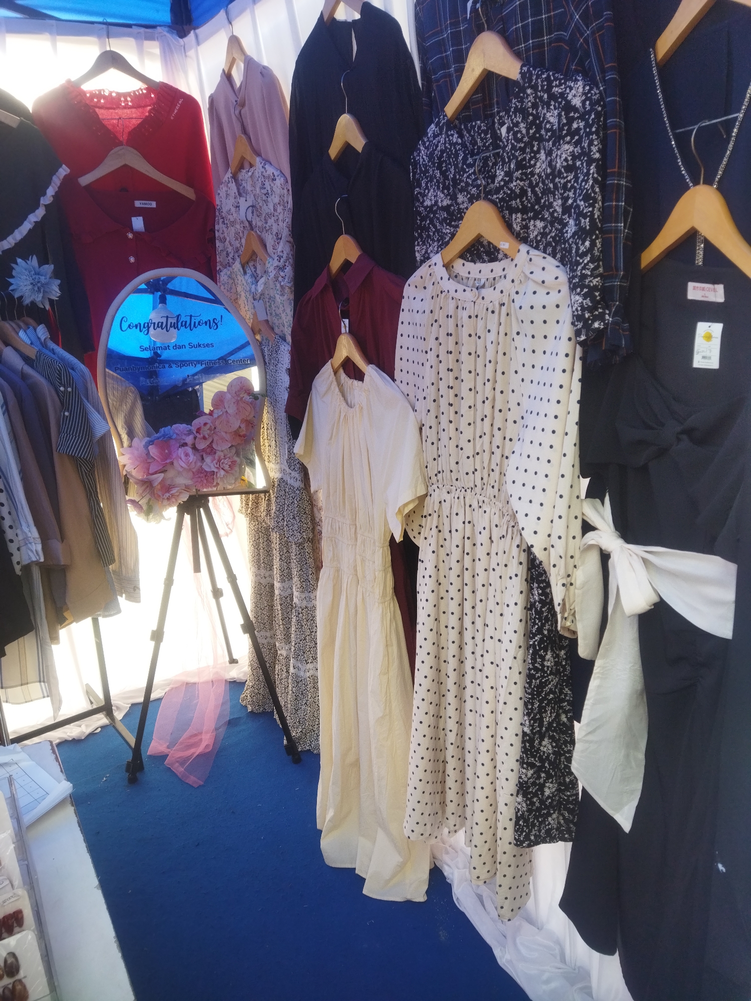 An outdoor fashion stall displays various garments for sale, including dresses and blouses, hanging on a rack under a blue canopy. A round mirror with a floral arrangement and text that reads "Congratulations! Selamat dan Sukses Puanbymonica & Sporty Fitness Center" is propped up on a tripod stand. The stall is situated on a blue carpet in what appears to be a marketplace, with other items and fabric drapes visible in the background. The lighting suggests it is daytime. The location is identified as Giripurwo, Indonesia.