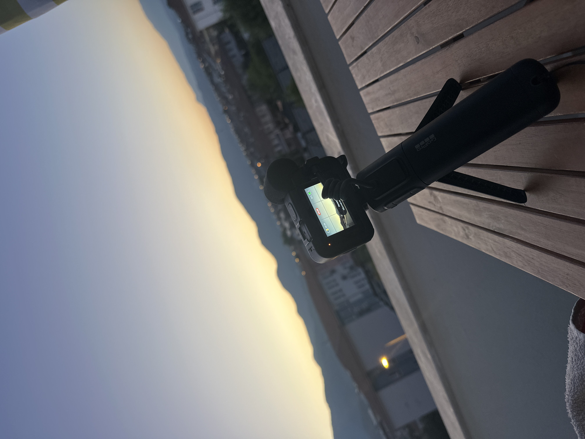 A GoPro camera is mounted on a tripod, positioned on a wooden outdoor table. The camera is capturing a sunset scene over a coastal city, likely Fethiye, Turkey. The sky displays a gradient of blue and orange hues, transitioning to darker tones as it meets the silhouette of distant mountains. The city below is illuminated by scattered lights, indicating dusk. The foreground shows the camera, tripod, and the wooden surface of the table. The overall environment suggests an outdoor balcony or terrace setting with a view of the cityscape and natural landscape at sunset.