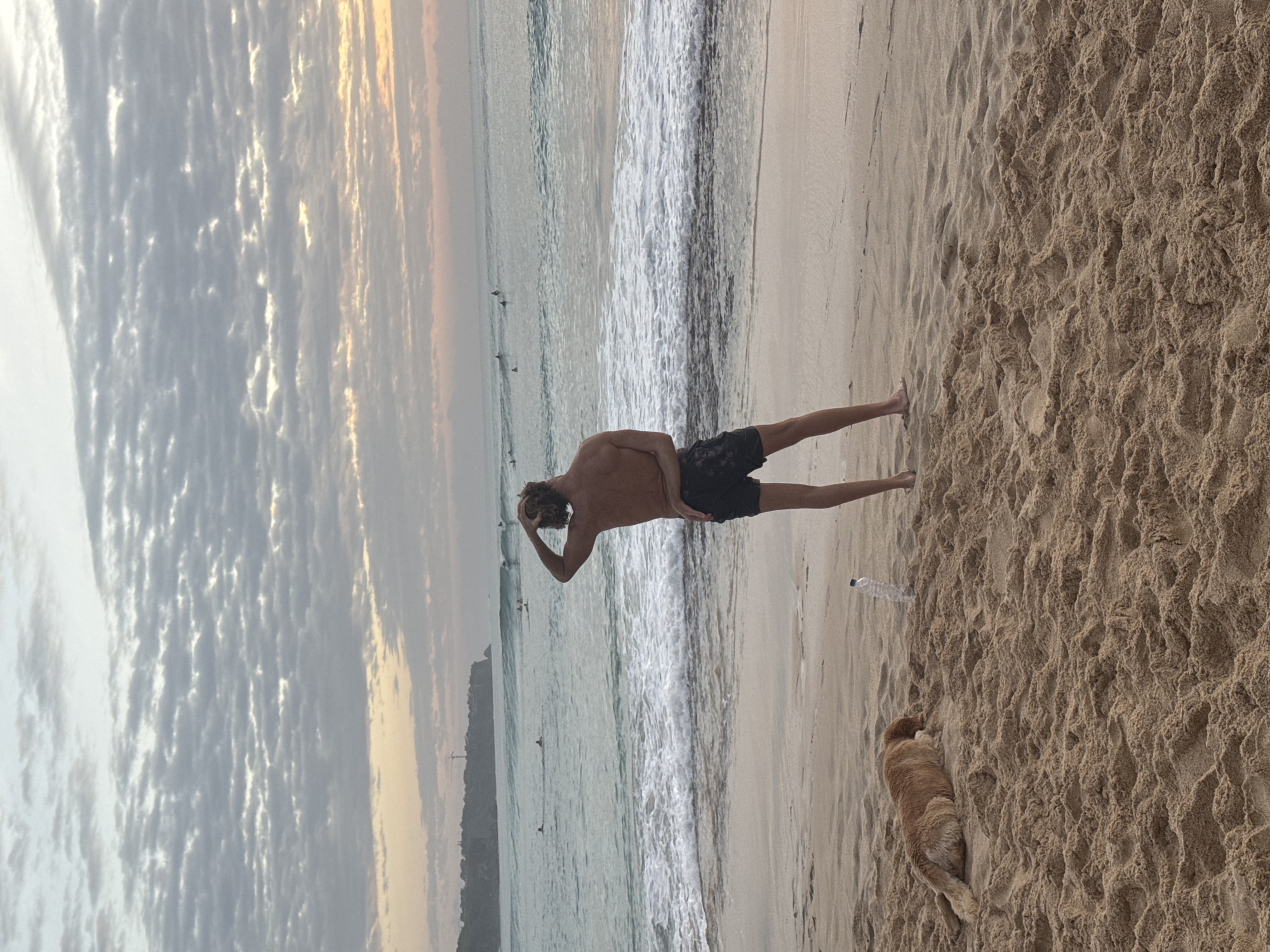 A wide shot captures a serene beach scene with a young man and a dog on the sand, facing the ocean. The man, with sandy blonde hair and wearing black swim trunks, stands with his back to the camera, one hand resting on his head. To his left, a golden-brown dog lies down on the sand, its head resting on its paws. A plastic water bottle stands upright in the sand between the man and the dog. The ocean features moderate waves and a group of surfers can be seen in the distance. The sky above is filled with a dramatic layer of textured clouds, tinged with the soft colors of dawn or dusk. In the far distance, a cliff face with a lighthouse or tower is visible. The beach is covered in light brown sand, with ripples and indentations indicating recent activity or wind.