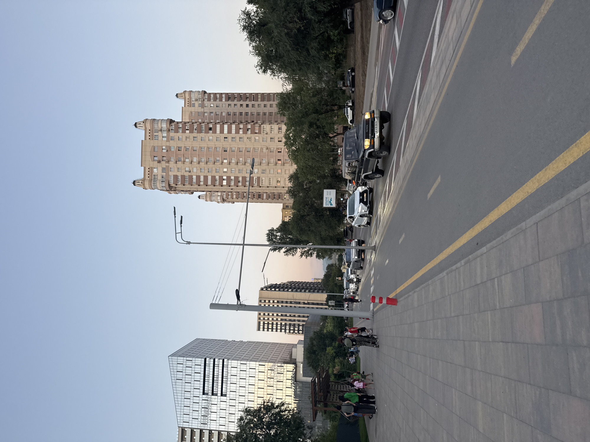 The image captures a street scene in Almaty, Kazakhstan, likely in the late afternoon or early evening, given the soft, diffused lighting. The foreground features a wide, paved sidewalk made of interlocking grey tiles. A few people are visible on the sidewalk, some walking, others standing or sitting near a covered shelter. Children are also present, some playing or interacting.
Adjacent to the sidewalk is a multi-lane road with clear markings, including yellow and white lines, and a dashed white line delineating lanes. Several vehicles are driving on the road. In the middle lane, a dark-colored SUV, possibly a Toyota Land Cruiser, is prominently featured, driving towards the viewer. Other cars, including a white Lexus and a dark sedan, are also on the road.
Dominating the background are several modern buildings. On the left, a tall, angular building with a glass facade reflects the sky and surrounding environment. To its right, a significantly taller, older-style residential building with a distinct beige and pink facade, featuring rounded balconies and ornate tops, is visible. Further back, other buildings and trees form part of the urban landscape. Streetlights and utility poles are also present along the roadside. The sky above is a clear, pale blue, transitioning to a warmer hue near the horizon, suggesting sunset. The overall impression is that of a busy urban street with a mix of modern and older architecture.