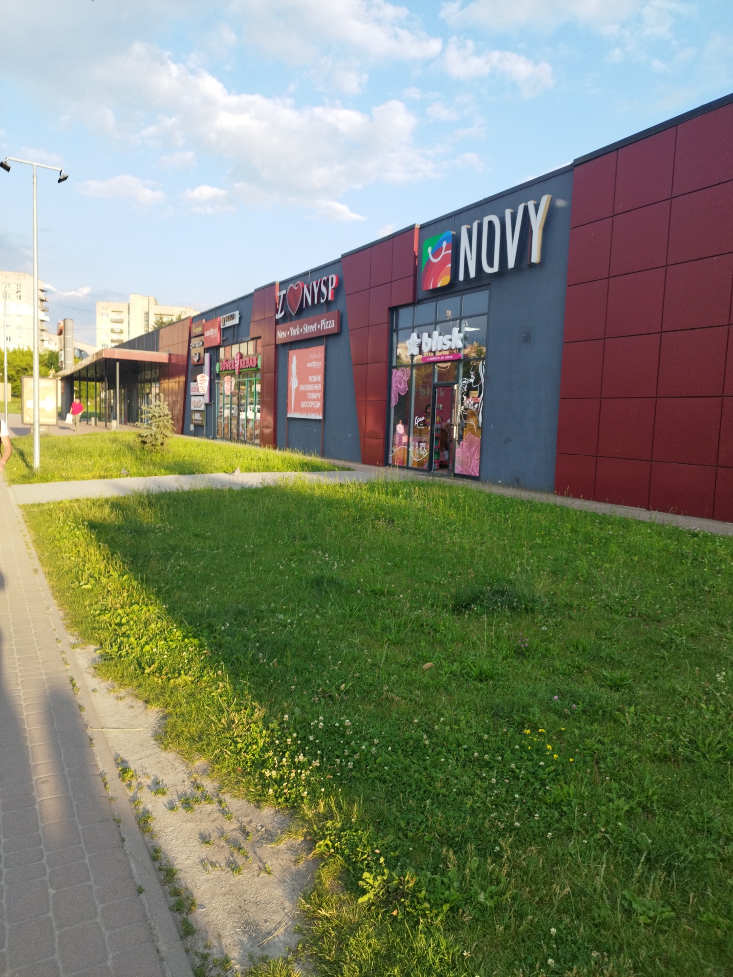 The image shows a street view of a commercial building with multiple storefronts in Novyi Rozdil, Ukraine. The building has a modern facade, with sections of dark gray and deep red cladding. Prominent signage includes "NOVY" in large white letters above a store entrance, and a "NYSP" logo with a heart design above another storefront. Several other shops are visible, including "DÖNER KEBAB" and "Blisk". The stores are situated along a paved walkway, bordered by a grassy area with scattered wildflowers. In the background, residential buildings and trees are visible under a bright blue sky with scattered clouds. The sun is casting long shadows, indicating it is late afternoon or early evening. There are no people clearly depicted engaging in any specific action, but one person in a red shirt is walking near an entrance. The overall scene is a typical urban commercial area on a clear day.