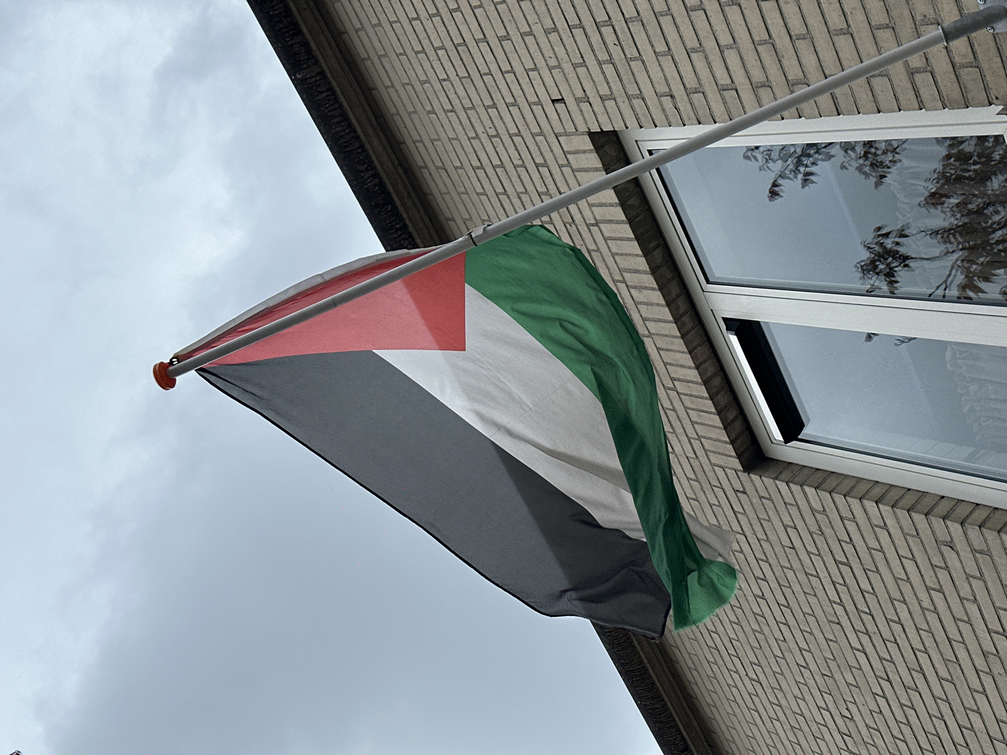 The photograph shows a Palestinian flag hanging from a pole attached to the exterior wall of a brick building. A window is visible on the wall, reflecting the sky and tree branches. The sky is overcast. The exact location is not specified and cannot be determined from the image.