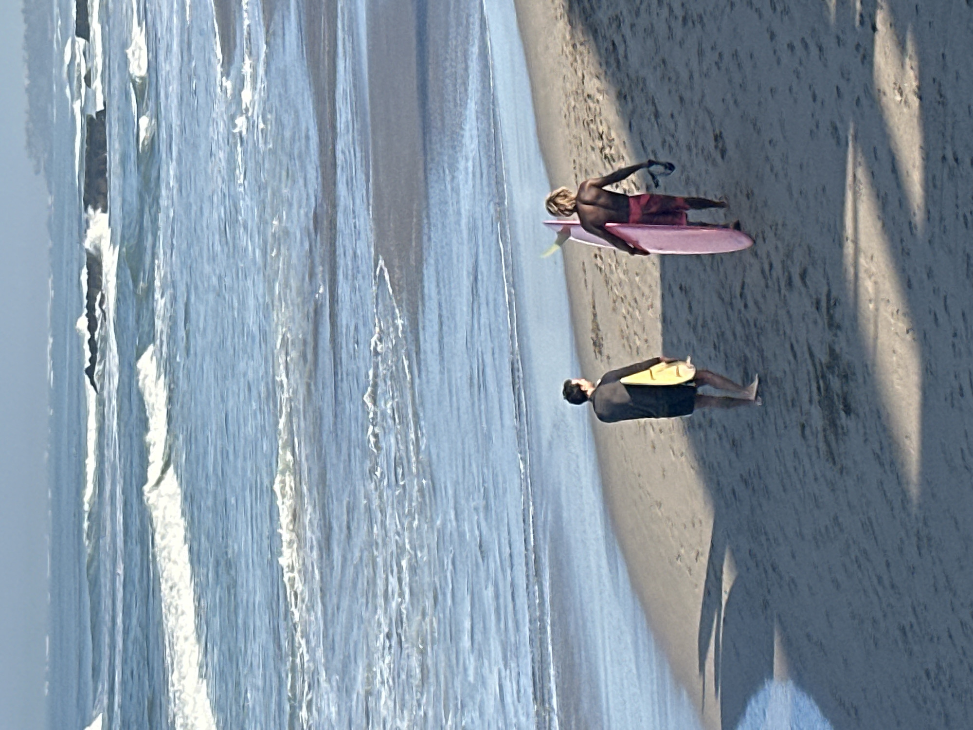 Two surfers walk along the sandy beach towards the ocean in North Kuta, Indonesia. The surfer on the left, wearing a black wetsuit top and shorts, carries a yellow surfboard. The surfer on the right, with blond hair and wearing red swim trunks, carries a pink surfboard and a pair of sandals. Gentle waves crash on the shore, and the sky is a soft blue with hints of clouds in the distance. The scene appears to be captured during the daytime, with the sun casting shadows on the sand.