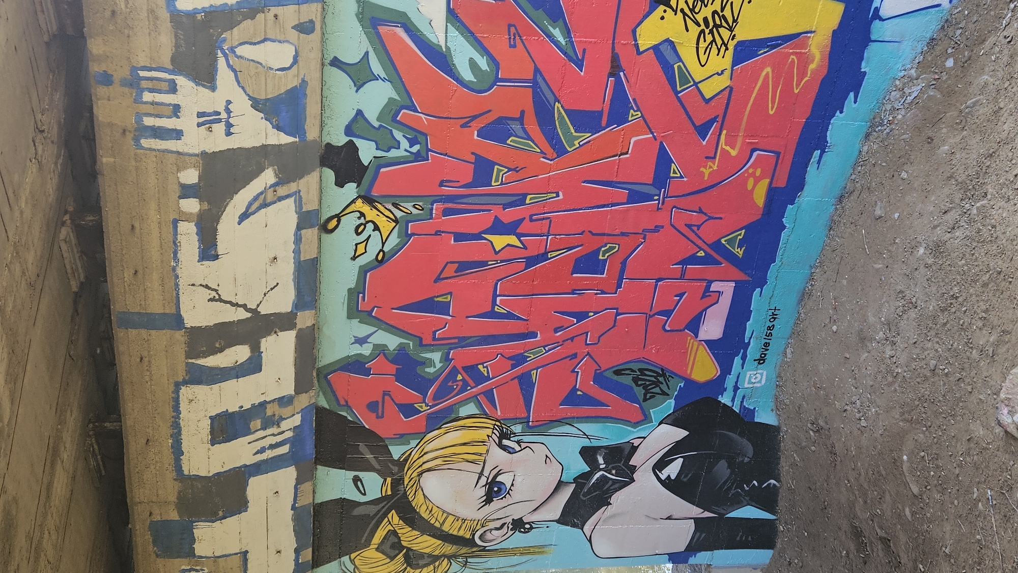 This is a photograph of graffiti art on the underside of a bridge or overpass. The graffiti is located in Prešov, Slovakia. On the left side of the image, there is a stylized anime-like illustration of a young woman with blonde hair tied in a ponytail with a black bow, large blue eyes, and a black outfit with a low neckline and a corset-like back. She is looking to the right, towards the graffiti. To the right of the illustration is a large, colorful graffiti piece featuring bold red lettering outlined in blue, with yellow accents and a small yellow crown on top. The background of the graffiti wall is painted teal. Above the main graffiti, there are abstract designs in white, blue, and gray. The ground in front of the wall is rough, sandy, and rocky. A small signature, "@dave158art", is visible in the lower right portion of the teal section.