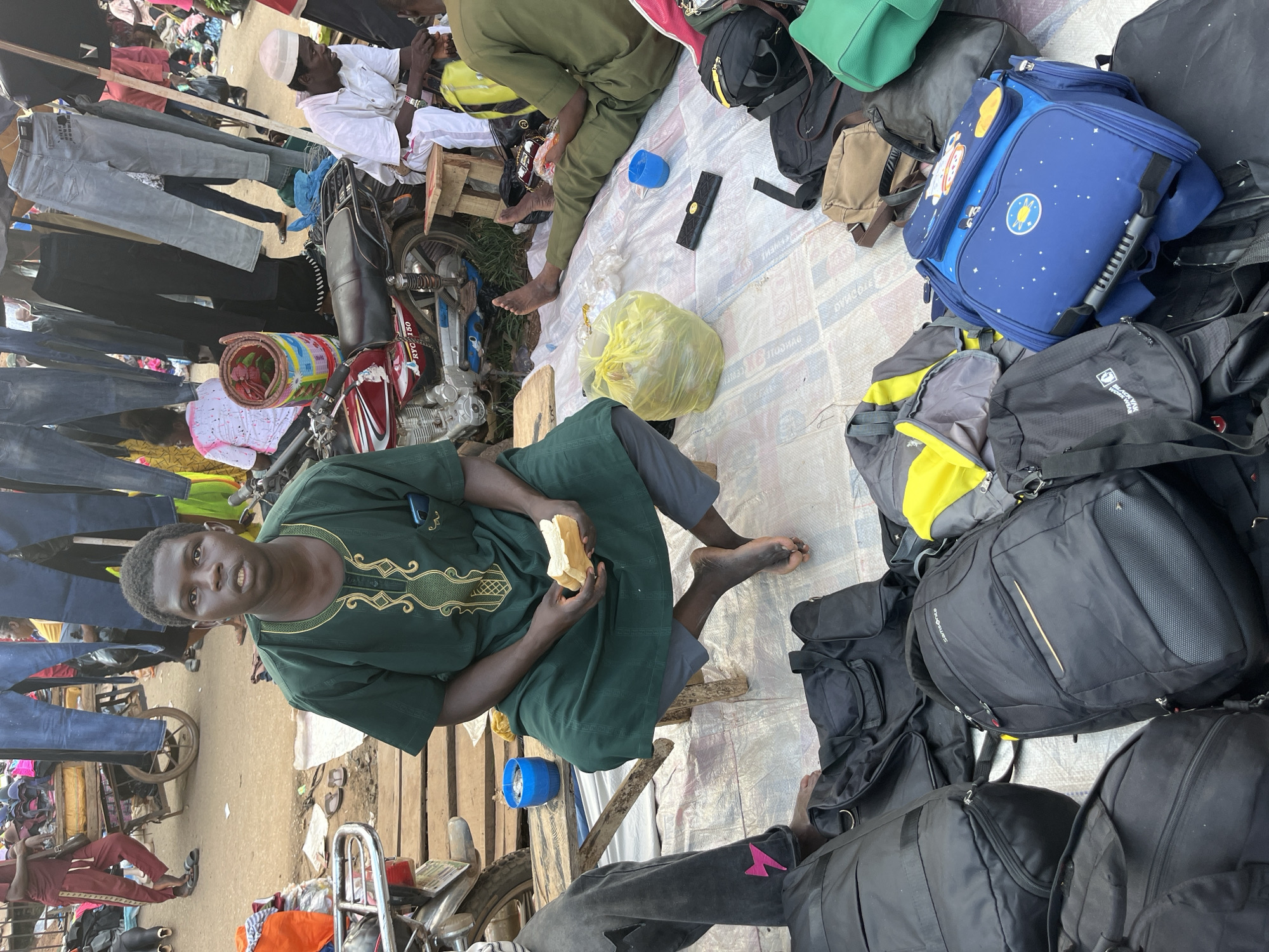 This image depicts a market scene in Nyanya, Nigeria. In the foreground, a young Black man is seated on a low wooden stool, wearing a green dashiki with gold embroidery. He is holding a piece of white bread in his hands. To his left, another person's legs are visible, wearing dark gray pants with a pink lightning bolt design.
The ground is covered with a patterned tarp, and a collection of bags, including large black duffel bags, a black Samsonite backpack, a blue child's backpack with space-themed decorations, and other assorted bags, are scattered around. To the left of the seated man, a blue plastic cup with some liquid and small objects inside is on a wooden plank. A red motorcycle is parked behind him.
In the background, the market is bustling with activity. Various stalls are visible, with clothing, particularly jeans, hanging from racks. People are seen browsing and interacting within the market environment. The overall scene suggests a vibrant, outdoor marketplace.