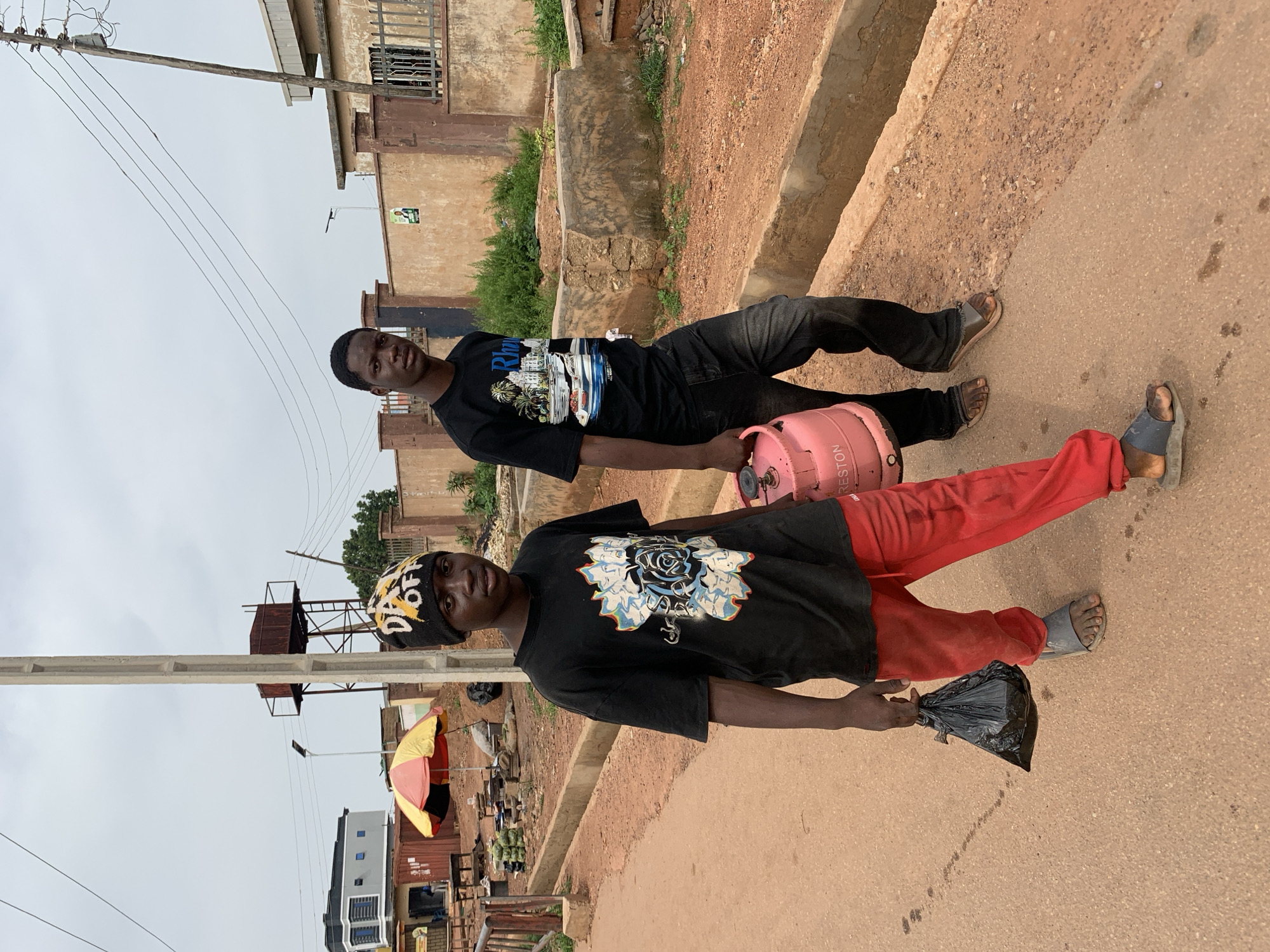 Two young men are walking on a dirt road in Jos, Nigeria. The man on the left is wearing a black t-shirt with a white and blue floral design, red pants, and gray sandals. He is carrying a black plastic bag in his left hand. The man on the right is wearing a black t-shirt with a design of buildings and palm trees, dark jeans, and brown sandals. He is carrying a pink gas cylinder in his right hand. Both men appear to be in their late teens or early twenties. The road is lined with concrete gutters and a few scattered buildings and utility poles. The sky is overcast, suggesting a cloudy day. The general environment appears to be a semi-urban or rural setting.