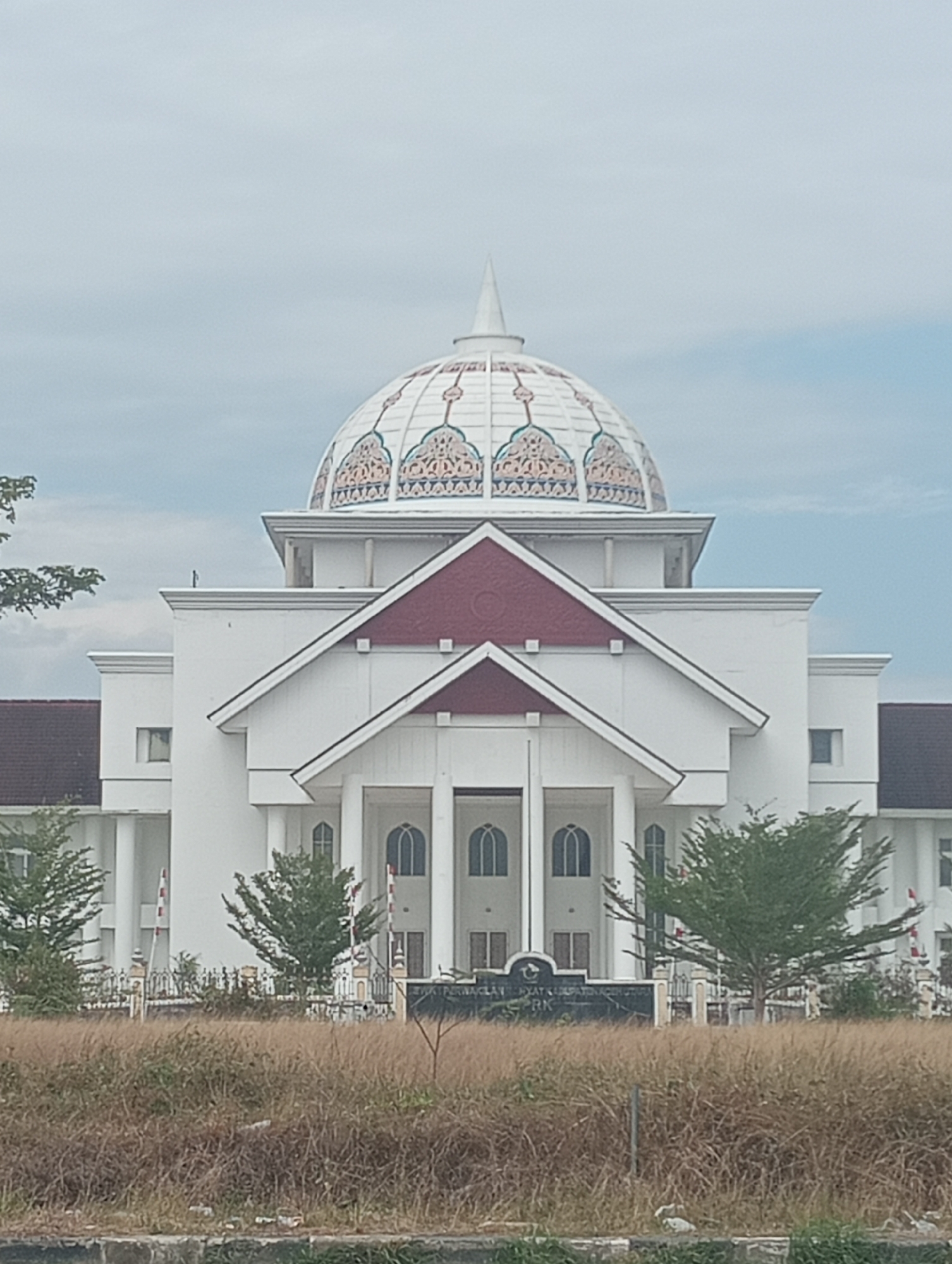 The image shows a white building with a distinctive domed roof. The dome is decorated with colorful patterns and a pointed finial. The building has a grand entrance with columns and arched windows. Red triangular accents are visible on the roofline. In front of the building, there is a sign with text that appears to be in Indonesian. The foreground is filled with dry grass and some sparse trees. The sky is overcast. The location is identified as Meunasahaluedrienlhoksukon Barat, Indonesia. There are no people visible in the image.