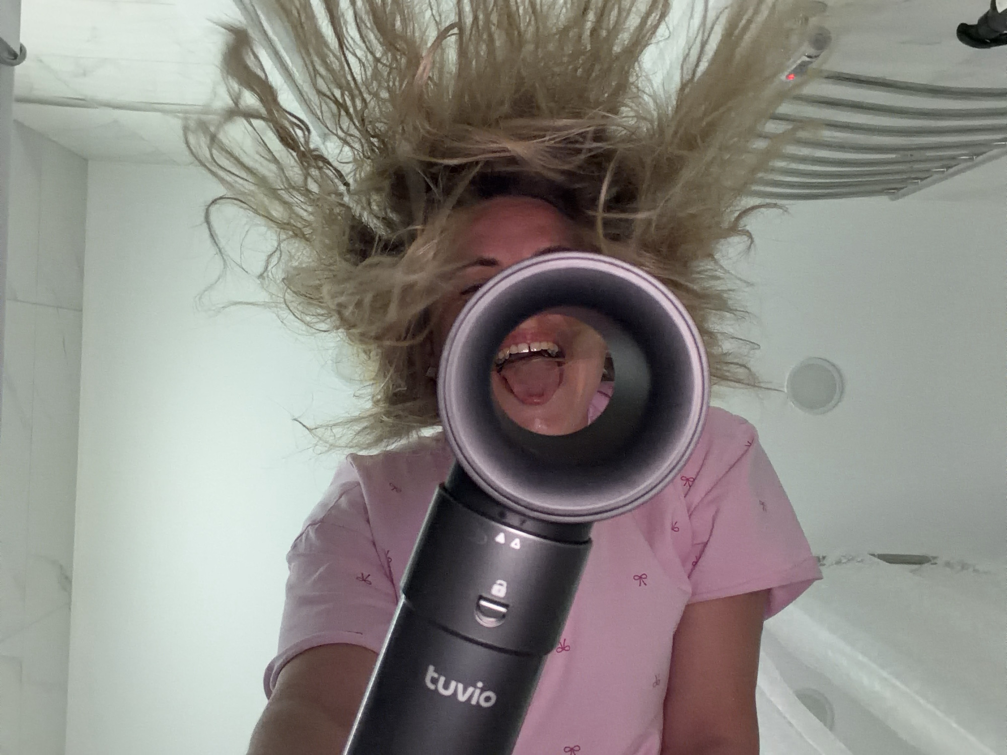 The image shows a low-angle perspective looking up at a person playfully interacting with a hair dryer in what appears to be a bathroom. The central subject is a person, likely female, whose face is partially visible behind a dark grey hair dryer pointed directly towards the viewer. Their light-colored, messy hair is dramatically blown upwards and outwards, creating a large, chaotic halo around their head, indicating the hair dryer is in active use.
The person's mouth is wide open, revealing their tongue and teeth, suggesting an expression of surprise, amusement, or the sensation of the air. They are wearing a light pink t-shirt with small, repetitive bow patterns printed on it.
The background consists of light-colored, possibly white, bathroom walls and ceiling. Visible bathroom features include a chrome-finished towel rack with horizontal bars on the upper right, and what looks like a white edge of a shower or bathtub enclosure on the lower right. Small circular fixtures, possibly ceiling lights or vents, are also visible.
The hair dryer prominently displays the brand name "tuvio" on its handle. Above the logo, there are various control symbols, including a lock icon and two arrow-like symbols, likely for operating the device. The image conveys a sense of lighthearted fun and experimentation.