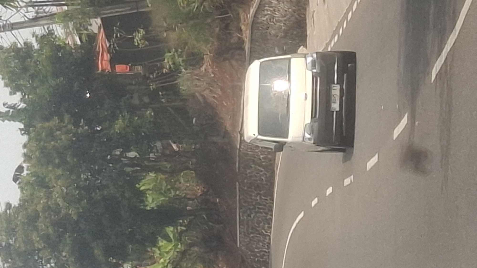 A light-colored van, likely a commercial vehicle, is driving on a paved road with white dashed lane markings. The van has a license plate that appears to read "B 885 TT". The road curves to the left, and a low stone wall lines the right side of the road. Beyond the wall, the terrain slopes upwards, covered in vegetation and trees. In the background, there are more trees and some buildings, including one with an orange roof. The sky is overcast. The image was taken in Sirnagalih, Indonesia, as indicated by the presence of a specific type of white van commonly used for transport in that region and the general appearance of the landscape consistent with rural Indonesia.