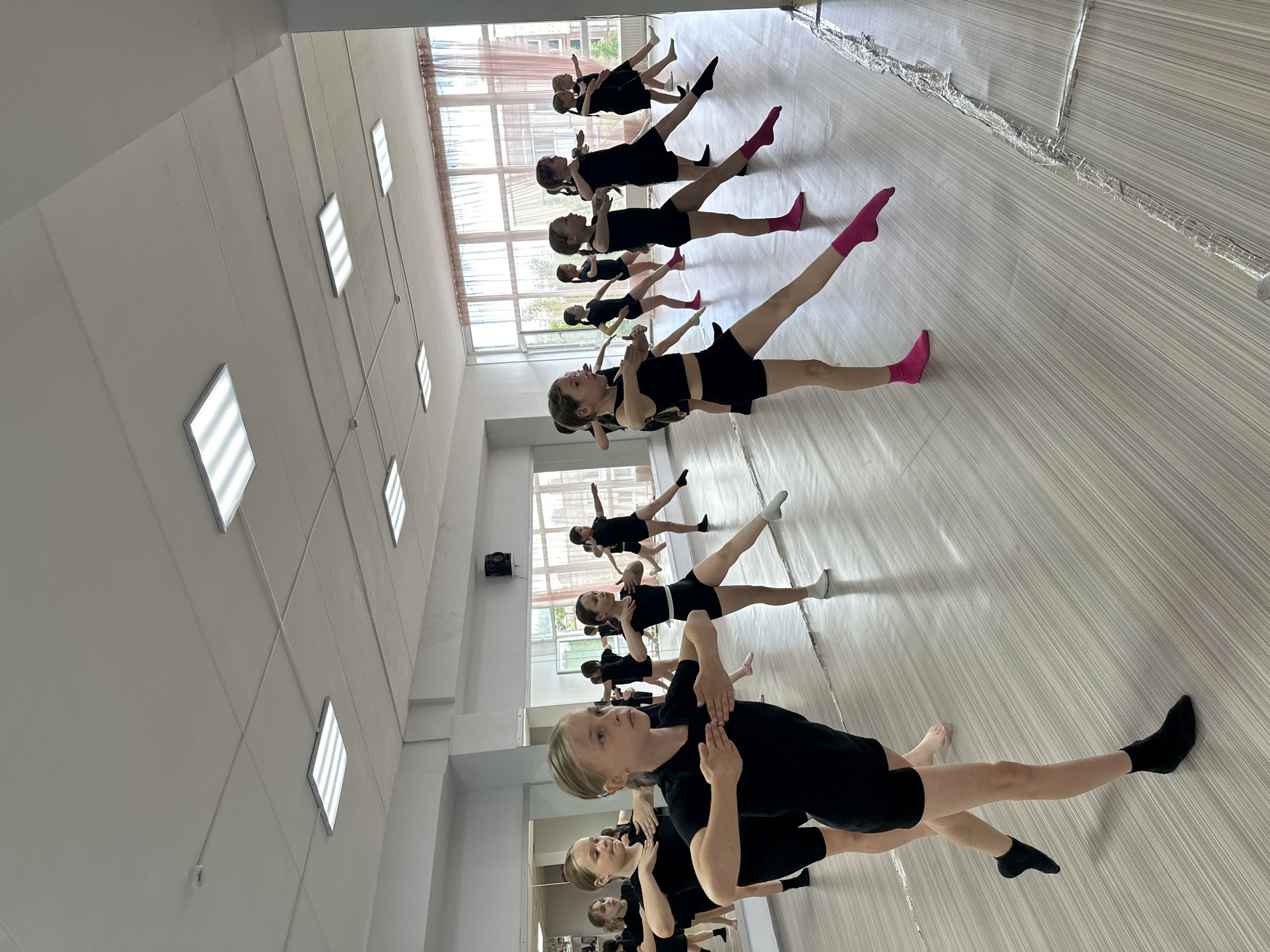 **Location:** Votkinsk, Russia.
**Scene Description:** Inside a dance studio. Approximately ten young girls are practicing a dance routine. They are positioned in rows, all wearing black leotards or similar attire. Some wear socks of various colors, including pink, white, and black. Their arms are held in front of their chests with their hands open, while one leg is extended to the side. The studio features a mirrored wall reflecting the dancers, and large windows providing natural light. Overhead, rows of square ceiling lights illuminate the space. The floor has a light-colored, striped pattern. There are support columns and walls painted in a light color.