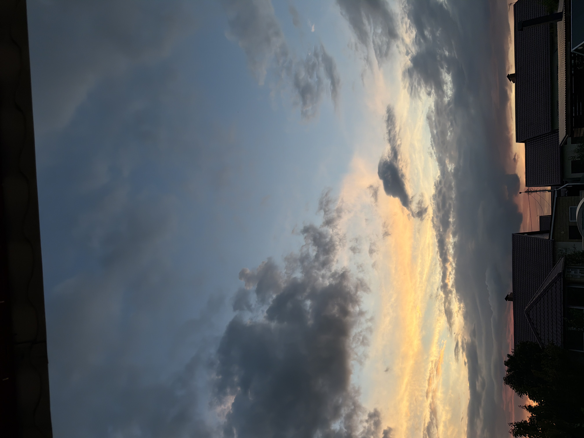 The image depicts a cloudy sky at dusk, with the sun setting and casting warm hues of orange and yellow across the horizon. Darker, heavier clouds are dispersed among lighter, wispy ones, creating a dramatic sky. Below the sky, the dark silhouettes of rooftops and trees are visible, suggesting a residential area. The scene is a typical view of a rural or suburban landscape at the end of the day. There are no people or actions to describe. The location is specified as Узюково, Russia.