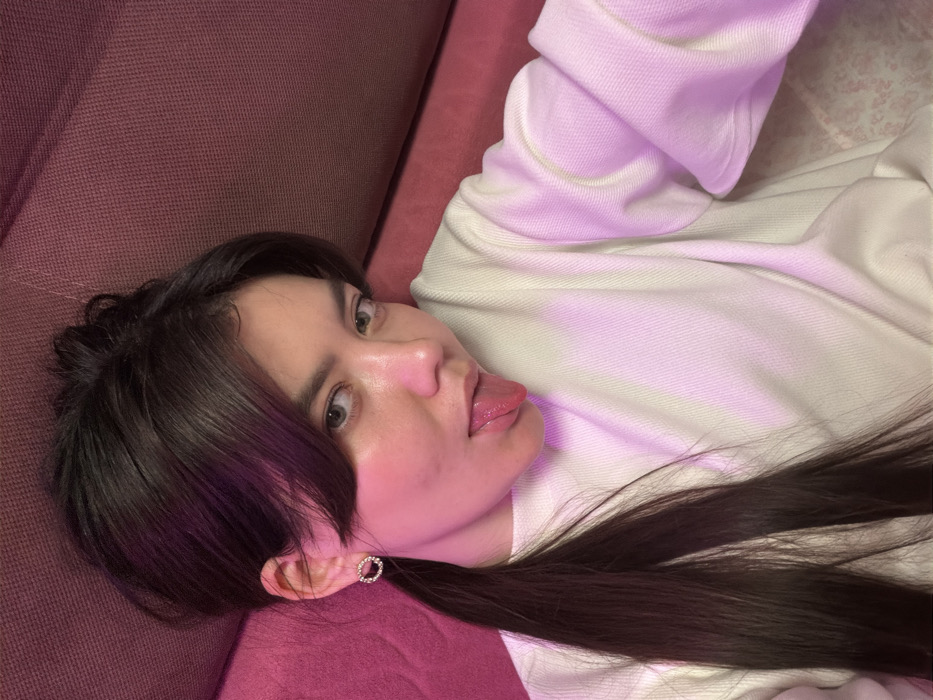 An image taken indoors depicts a young female posing, likely for a selfie, in the city of Chaikovsky, Russia. The subject, exhibiting fair skin, dark brown hair styled in a high ponytail with bangs, and light-colored eyes, is seen from the chest up. She wears a plain white long-sleeved top, possibly a ribbed sweatshirt, and a small, circular, gold-toned earring on her visible left ear. Her head is tilted slightly, and she is looking directly at the camera with her tongue extended. The background consists of a textured, dark pink or magenta upholstered surface, possibly a sofa or bed. Ambient pink-purple light is visible on the subject's face and clothing, particularly on her right side. No other individuals or distinguishing objects are present in the frame.