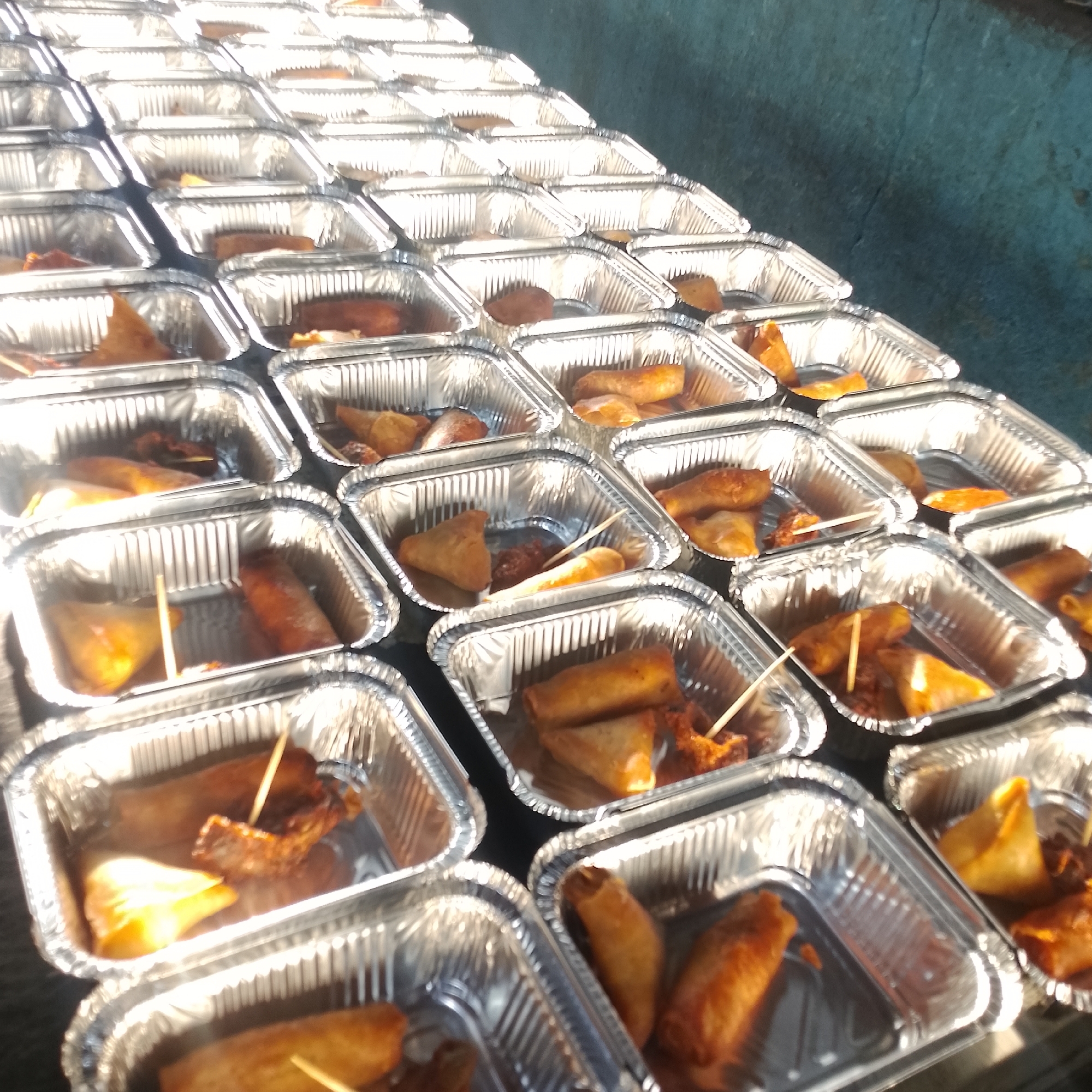 In Lagos, Nigeria, rows of foil food containers are neatly arranged on a dark, textured surface, likely asphalt or concrete. The containers are filled with a variety of fried foods, including samosas, spring rolls, and what appear to be pieces of fried chicken or fish. Some items are skewered with toothpicks. The lighting suggests an outdoor setting, possibly a market or event, with sunlight casting reflections on the foil. The overall scene depicts a food distribution or catering setup.