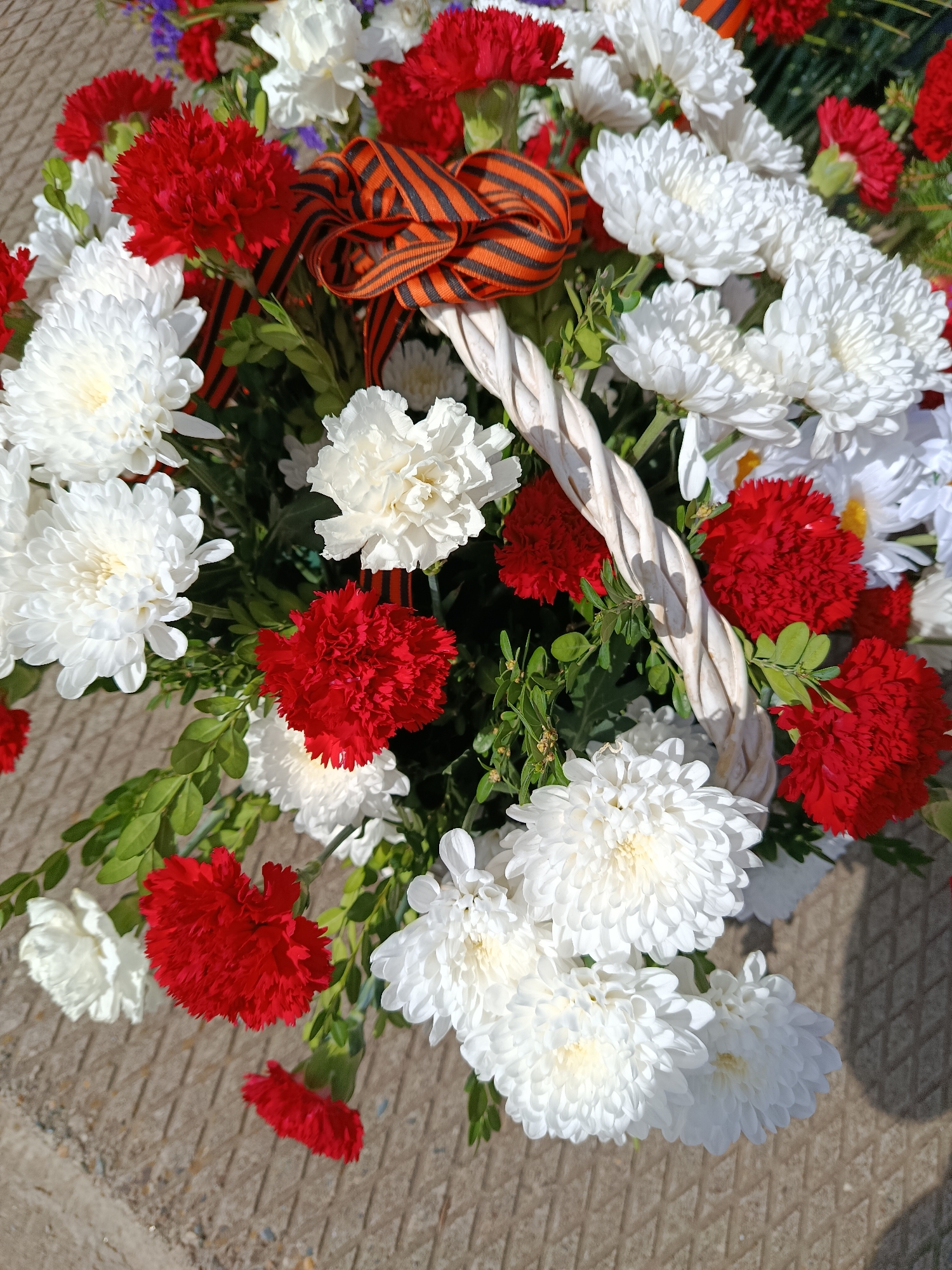 A close-up, eye-level shot depicts a flower arrangement in a white wicker basket placed on a concrete surface. The basket contains a mix of red carnations and white chrysanthemums, interspersed with greenery. An orange and black striped ribbon is nestled among the flowers. The image is likely taken in Фоки, Russia.