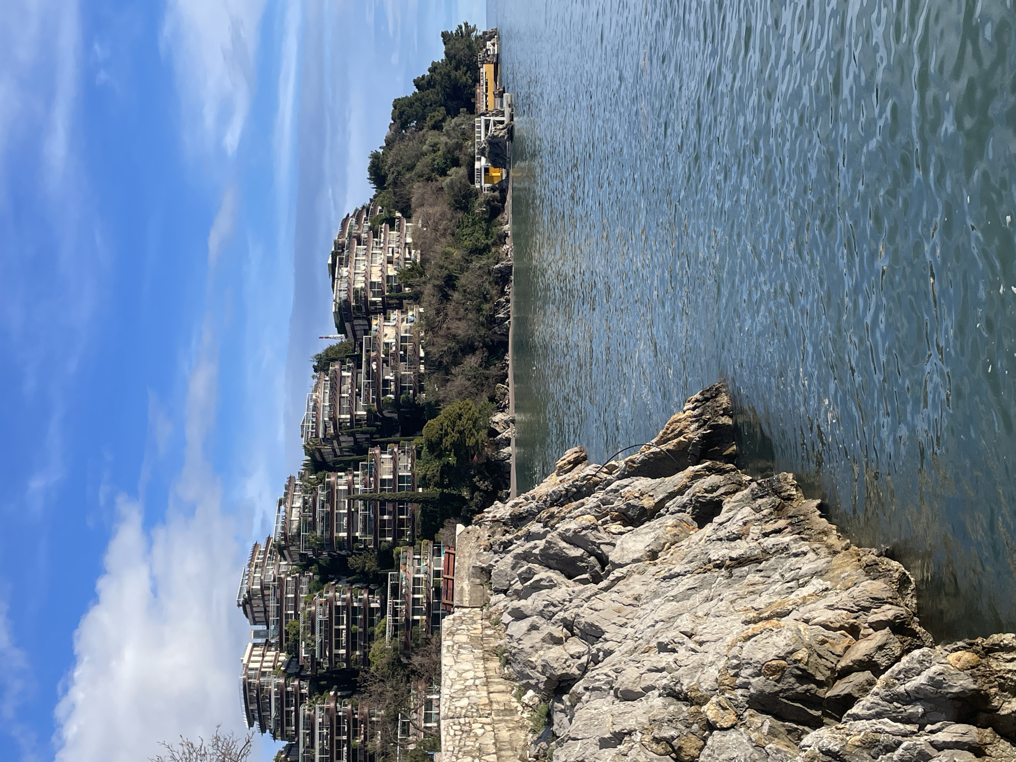 The image depicts a coastal scene in Budva, Montenegro, under a clear blue sky with scattered white clouds. In the immediate foreground on the left, a large, rugged rock formation, predominantly grey with some ochre tones, extends into the calm, blue-green sea. Above these rocks, a low stone wall, constructed from irregularly shaped light-colored blocks, runs horizontally.
Across the midground, the sea exhibits gentle ripples. Along the distant shoreline, a narrow stretch of light-colored sand is visible, backed by a dense line of trees and other vegetation on the hillside. Further along the coast to the right, several low-rise structures, prominently featuring yellow-colored facades, are situated close to the water's edge, possibly extending over piers.
The background is dominated by a steep, verdant hillside densely covered with numerous multi-story buildings. These structures are terraced, featuring light-colored facades, multiple levels of balconies with dark railings, and flat roofs. The buildings are interspersed with lush green trees, including tall cypress-like trees. No individuals or discernible activities are present within the frame.
