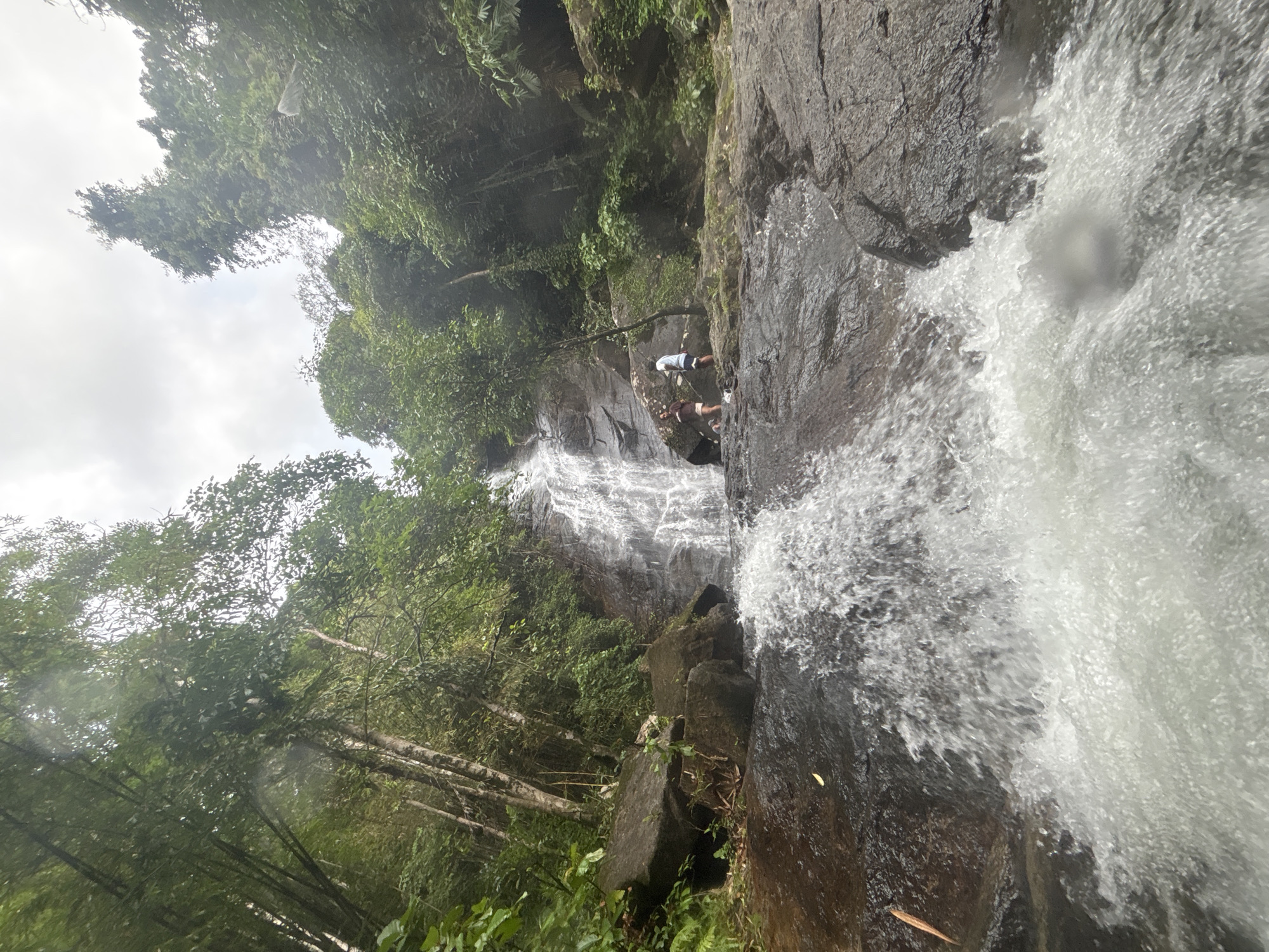 The image depicts a waterfall in Ban Bang Khli, Thailand. Two individuals are positioned on the rocky cliff face behind the waterfall. The surrounding environment is lush with trees and vegetation. The sky is overcast. Water is cascading down the rocks, creating a strong flow.
