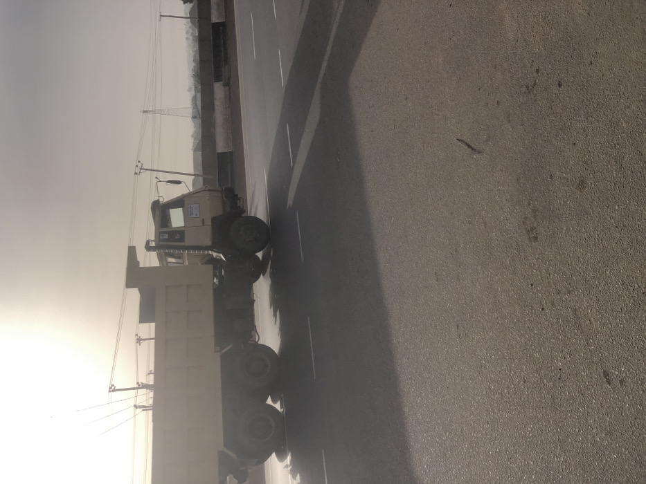 A dump truck sits on the roadside beside a paved road. Overhead power lines extend into the distance. The sky is overcast. Trees and vegetation are visible in the background near a roadside barrier. The road surface is asphalt.