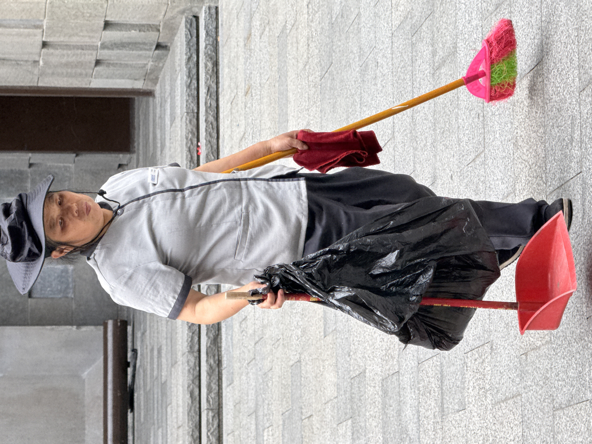 In the Yuzhong District of China, a person wearing a dark wide-brimmed hat and a grey uniform with dark trim is holding a black plastic trash bag filled with items. The trash bag is attached to a red dustpan, which is held at the bottom. The person is also holding a red cloth in their left hand and a broom with a yellow handle and a pink and green brush head in their right hand. The broom is positioned low to the ground, as if in use. The individual is standing on a paved outdoor area with large stone steps in the background. The overall environment suggests a street or public space within a city.