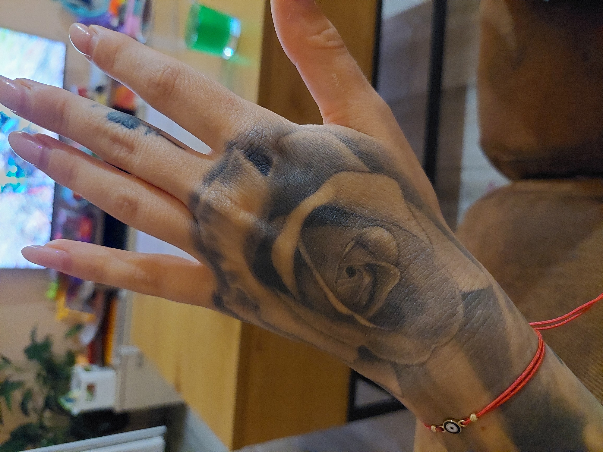 The image shows a close-up of a person's left hand, which is tattooed with a rose design that covers a significant portion of the palm and extends onto the fingers. The hand also has a red string bracelet with a blue and white evil eye charm on the wrist. The background is blurred but appears to be an indoor setting with a wooden table and a television screen in the distance. There is no visible information or context that places this image in Plovdiv, Bulgaria.