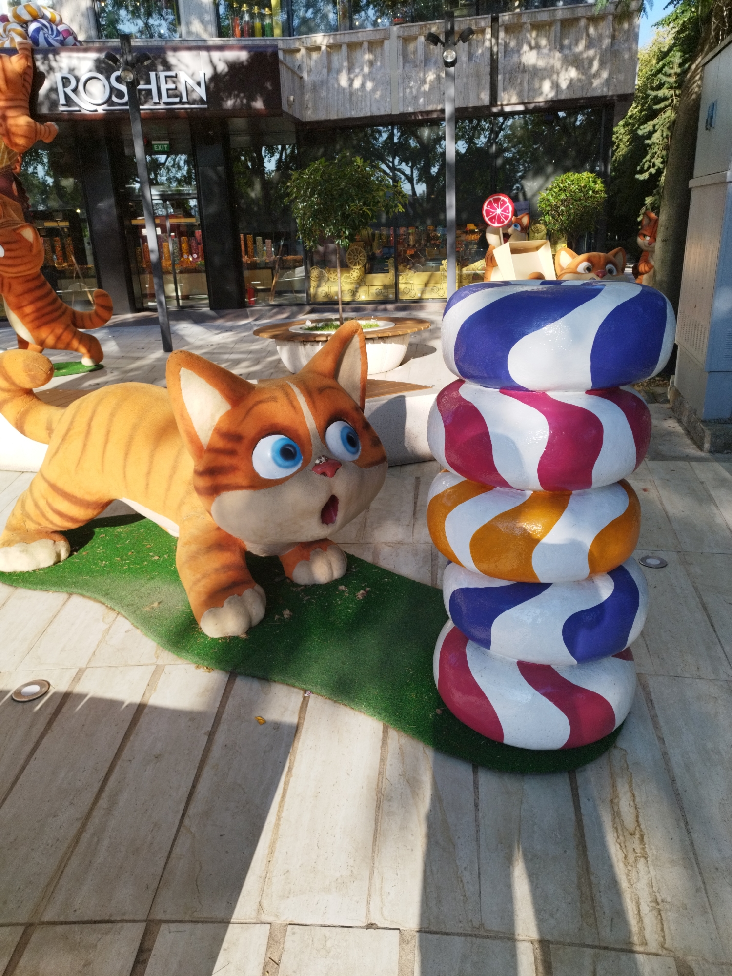 The image shows a playful outdoor display in front of a Roshen confectionery store, a well-known brand with a presence in Bucharest, Romania. In the foreground, a large, cartoonish orange tabby cat sculpture is posed on a patch of green artificial grass, with its mouth open in a surprised expression and blue eyes wide. To the right of the cat, a stack of five large, colorful, donut-shaped sculptures are arranged vertically. Each donut features a swirling pattern of two contrasting colors: blue and white, pink and white, orange and white, blue and white, and pink and white from top to bottom. In the background, the facade of the Roshen store is visible, with large windows displaying rows of candies and a prominent "ROSHEN" logo. Other decorative elements include small animal sculptures, one holding a lollipop, and trees. The ground is paved with light-colored tiles, and shadows from the sculptures and overhead structures are cast across the scene. The overall impression is one of a whimsical and inviting retail environment.