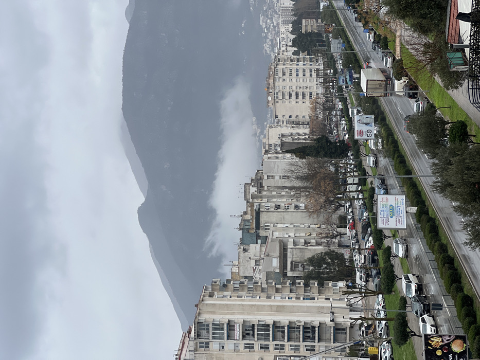 The image displays an urban landscape in Bar, Montenegro, on an overcast day with visible wet surfaces indicating recent rain. In the background, dark, rugged mountains are partially enveloped by a dense layer of white clouds or fog, obscuring their lower slopes and peaks. The midground features a dense array of multi-story residential buildings, predominantly light-colored with balconies and visible rooftop antennas, extending across the frame and receding towards the base of the mountains.
The foreground is dominated by a wet, multi-lane asphalt road running diagonally, appearing to be a major thoroughfare. Vehicles, including multiple cars, vans, and a white semi-truck, are present on the lanes and in numerous parallel parking spaces along both sides of the road. A central median strip, planted with green hedges and bare deciduous trees, separates the traffic directions. Streetlights are spaced along the road. Several billboards are visible: one on the median displays "MIX" with Cyrillic text, another shows "50," and a third on the left advertises food items. Green shrubbery and trees are present along the road margins and in the immediate foreground. No specific individuals are discernible within the scene.