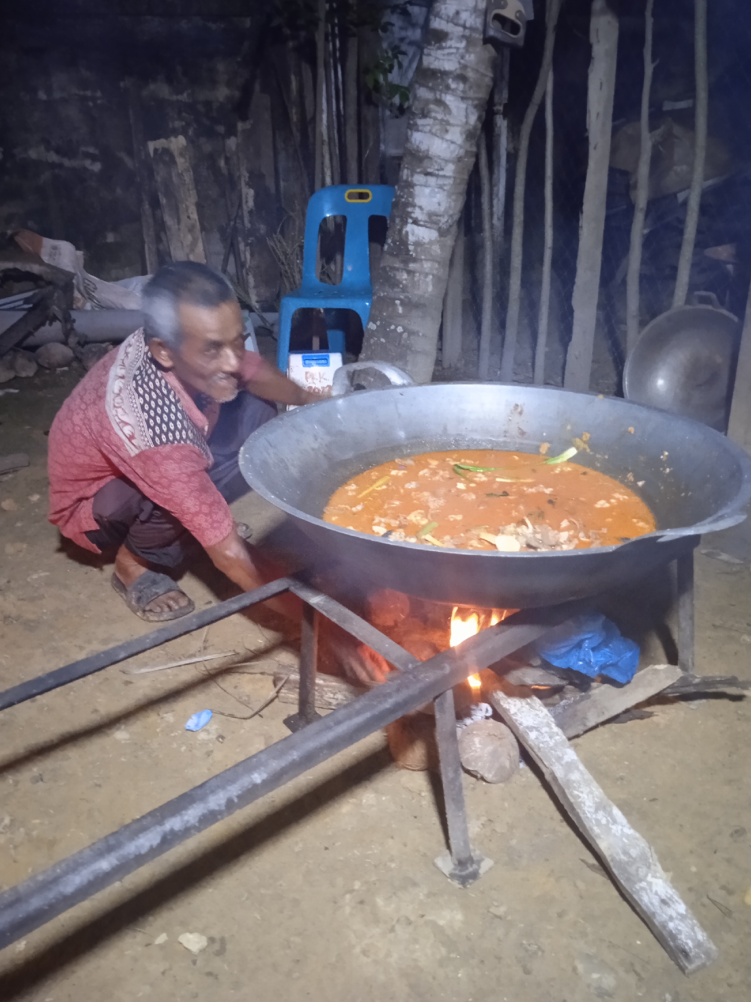 An elderly man with gray hair and a mustache is squatting down in front of a large metal pot over a fire. He is wearing a reddish-pink patterned long-sleeved shirt and dark pants, with dark sandals on his feet. The pot is filled with a reddish-orange stew with pieces of meat and green vegetables. The fire below is burning brightly, with flames visible. The scene appears to be outdoors, possibly in a village setting, with wooden poles and trees in the background, and a blue plastic chair to the right. The ground is made of dirt. The overall atmosphere is rustic and suggests outdoor cooking. There is no specific information to determine the location within the city of Keudeteupinpunti, Indonesia, from the image alone.