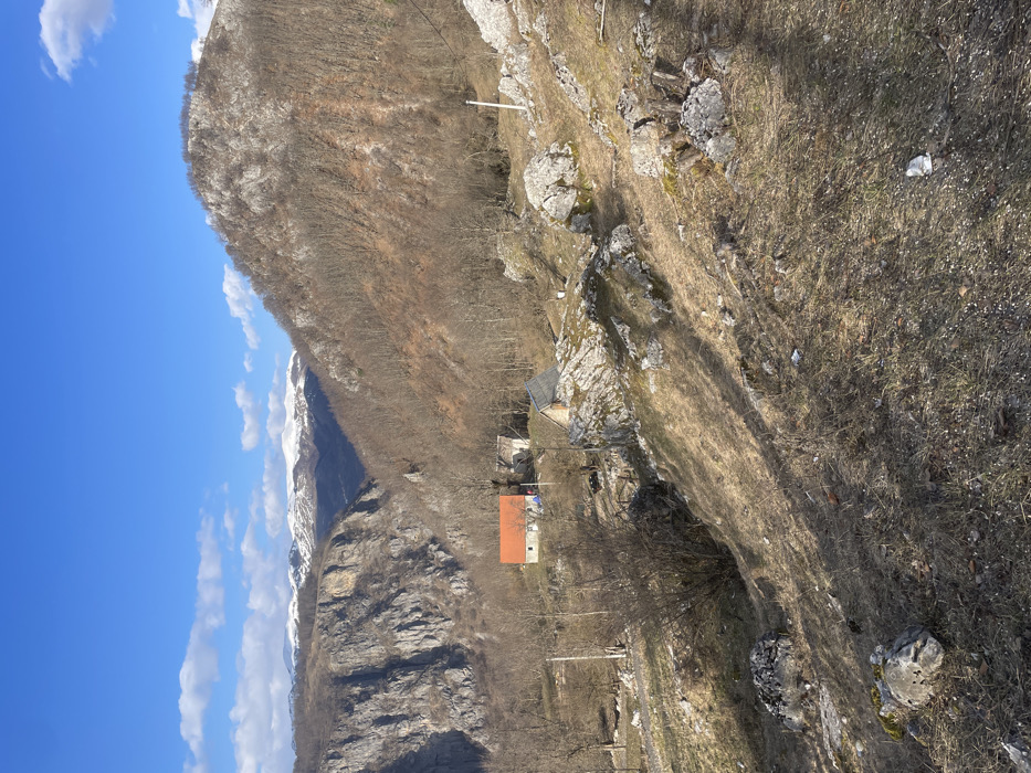 The image depicts a mountainous rural landscape in Mokro, Montenegro, under a clear blue sky with scattered white clouds. The scene is presented from an elevated viewpoint looking down into a valley.
In the immediate foreground, a dry, sloping terrain is visible, characterized by brown and pale green dry grass, exposed soil, and numerous light gray rocks and boulders. Sparse, leafless bushes and tree stumps are interspersed throughout this area. A piece of white crumpled debris is visible on the ground towards the lower right.
The midground features a small cluster of structures situated within a more level, partially vegetated area at the base of the slope. The most prominent building is a light-colored house with a distinct orange-red gabled roof. Adjacent to it on the right is another light-colored building, possibly a barn or an older dwelling, with a partially visible dark roof. To the right and slightly in front of these two buildings, a smaller, darker structure with a dark roof and lighter walls is present, appearing to be a shed or outbuilding. Utility poles with overhead lines are visible in the midground, with bare deciduous trees and bushes surrounding the structures and extending up the slopes to the right.
The background reveals a deep canyon or valley. On the left, a sheer, rocky cliff face rises dramatically. On the right, a densely forested mountain slope, primarily consisting of bare deciduous trees, ascends. In the distant background, a range of snow-capped mountains is visible beneath the horizon. There are no individuals or discernible actions occurring within the frame. The lighting suggests a bright, sunny day.