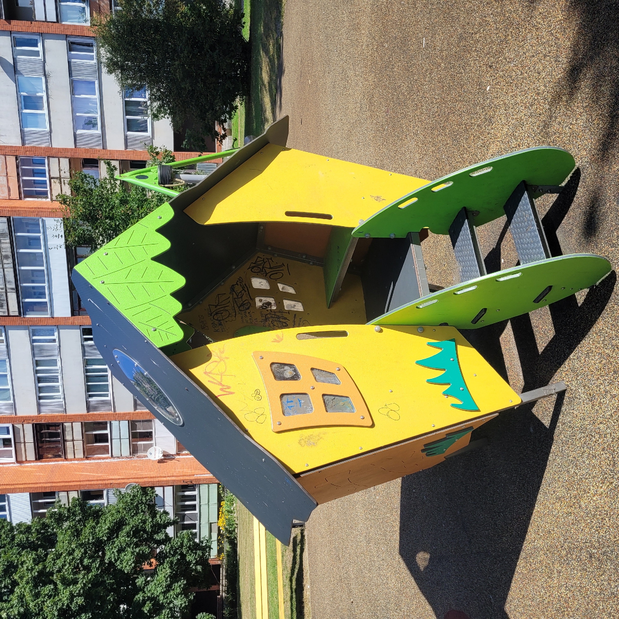 A bright yellow, multi-sided playhouse with a grey and green roof sits on a textured, brown surface, likely a playground. The playhouse has a circular window on the roof and a square window with four panes on one side. Green steps with grey metal treads lead up to an open doorway. The interior of the playhouse is visible, with a green bench and dark grey flooring. The exterior walls of the playhouse are decorated with simple green leaf shapes and some graffiti. In the background, a large brick apartment building with many windows is visible, along with trees and a yellow bench. The scene is bathed in sunlight, casting shadows on the ground. The location is identified as Jurmala, Latvia. There are no people in the image.