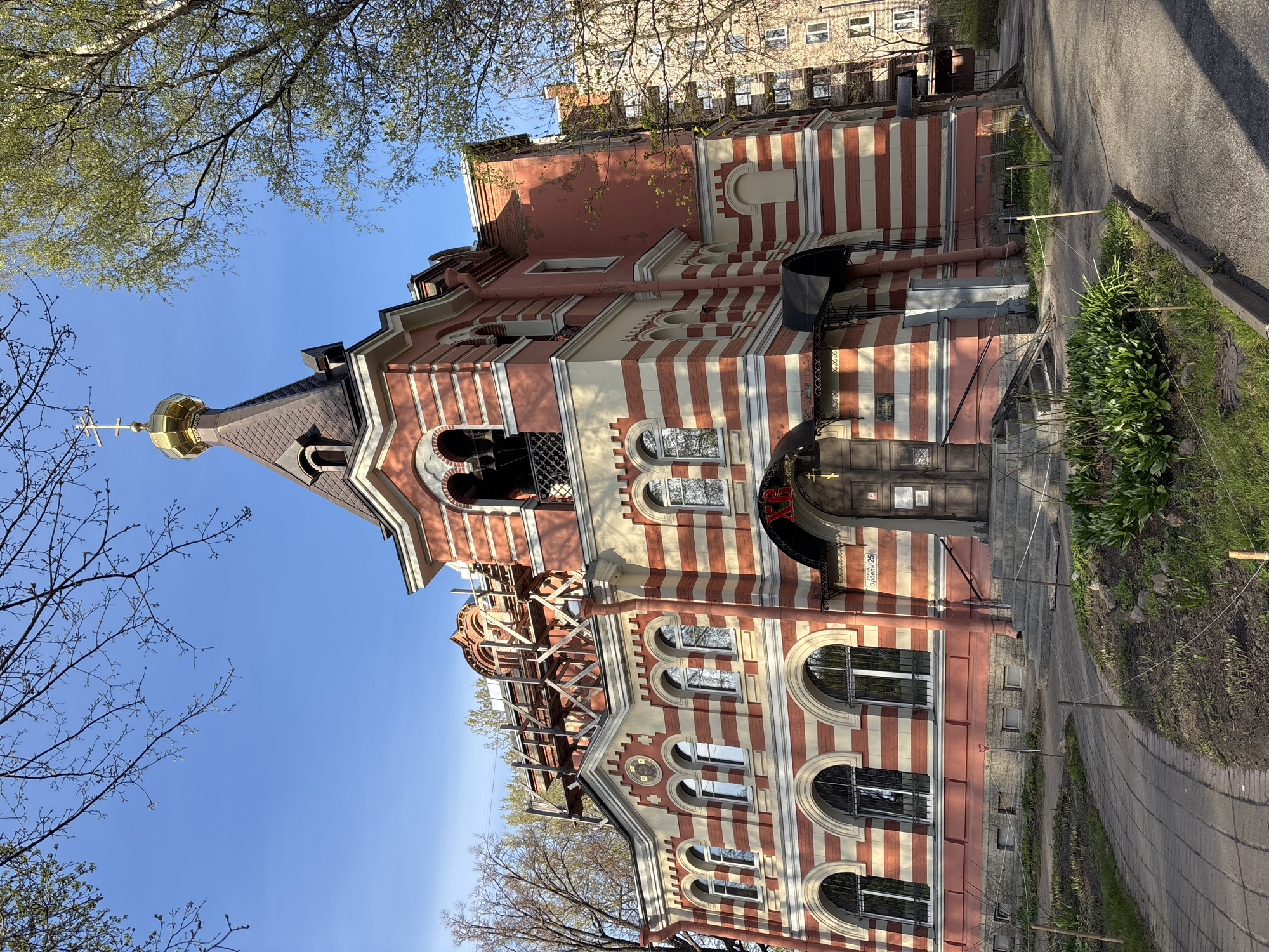 The image shows the Church of the Assumption of the Blessed Virgin Mary, a Russian Orthodox church located on Lieutenant Schmidt Embankment 27/2 in Saint Petersburg, Russia. The building is characterized by red and white stripes on its exterior walls. It has a black roof topped with a golden onion dome and a cross. Some scaffolding is visible on the side of the church. The sky is clear and blue. The church is surrounded by trees, some of which are bare, while others have new leaves. A sidewalk and a small patch of greenery are visible in the foreground. The image is taken from a low angle, looking up at the church.