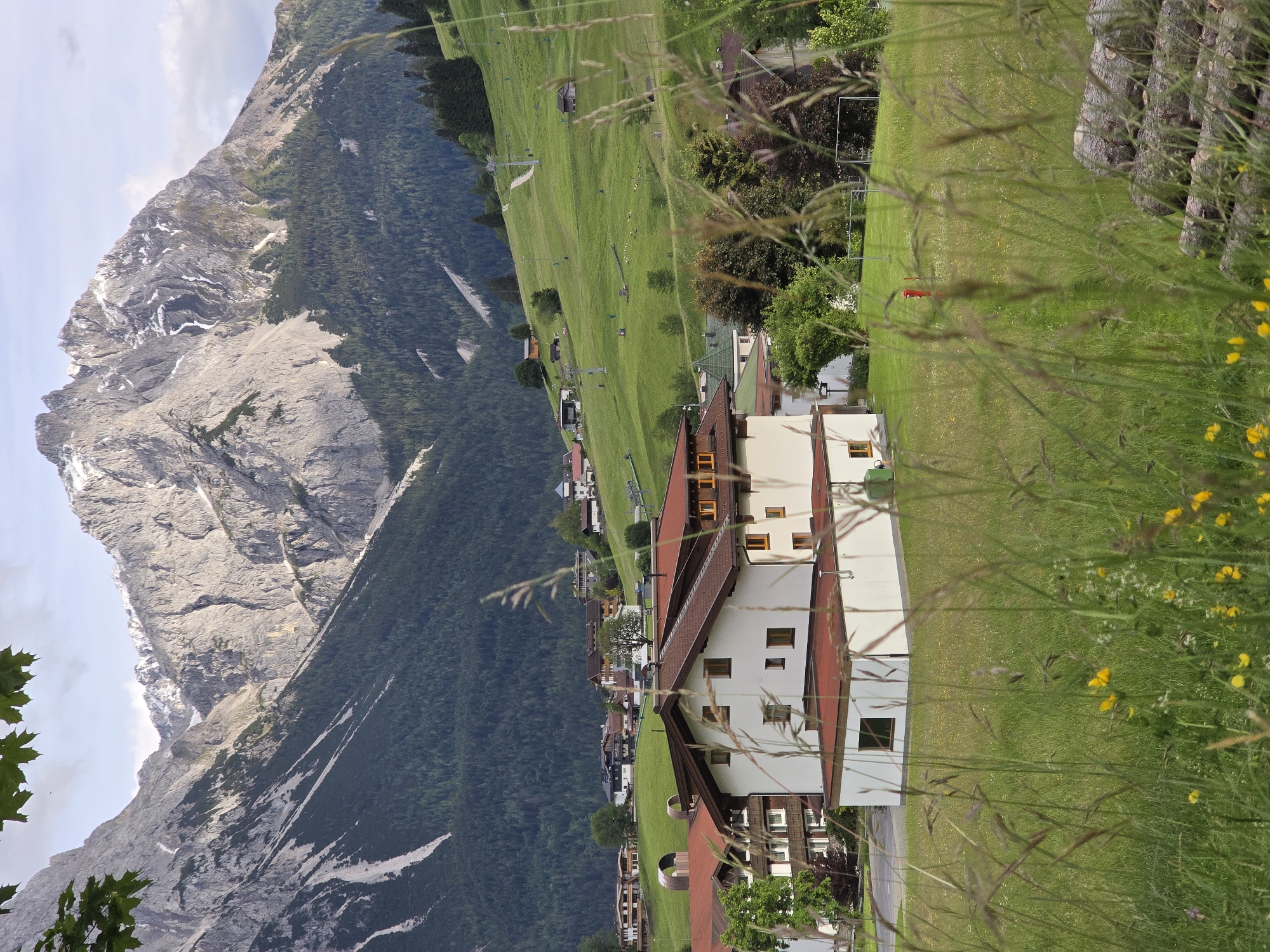 This is a vertical, eye-level shot of a scenic view in Lermoos, Austria, captured on a bright, partly cloudy day. The foreground is dominated by a lush, green meadow with tall grasses and scattered yellow wildflowers. In the midground, a large, white building with a reddish-brown roof stands prominently, featuring numerous windows and balconies. To the left and right of this building, other structures with similar architectural styles are visible, suggesting a village or resort setting.
The background is a dramatic panorama of the Tyrolean Alps. Jagged, grey mountain peaks, some capped with snow, rise majestically against a pale blue sky with scattered white clouds. The mountainsides are covered in dense green forests, with a ski lift visible ascending one of the grassy slopes in the mid-distance. A few smaller buildings are scattered across the lower slopes of the mountains. The overall impression is one of a tranquil alpine village nestled beneath imposing natural beauty. No people or animals are discernible in the image.