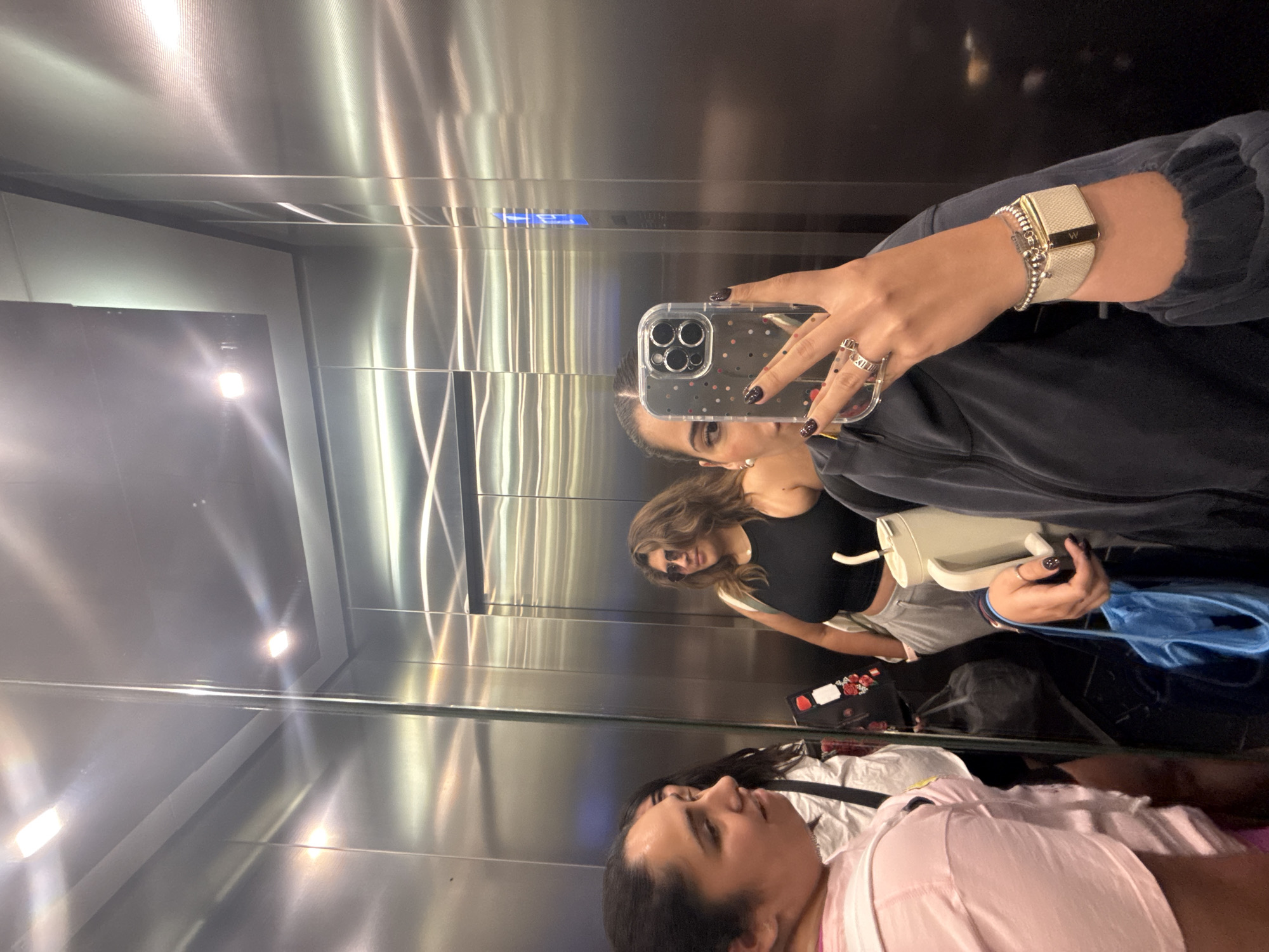 The image shows a reflection in an elevator. Three women are visible in the reflection. One woman holds a smartphone with a clear case and black nail polish, taking the picture. She wears a watch and rings. A second woman stands behind her, wearing sunglasses and a black top. A third woman is in the bottom of the reflection wearing a pink shirt. Luggage and a bag are also visible. The location is likely Puebla, Mexico, but this cannot be confirmed from the image.
