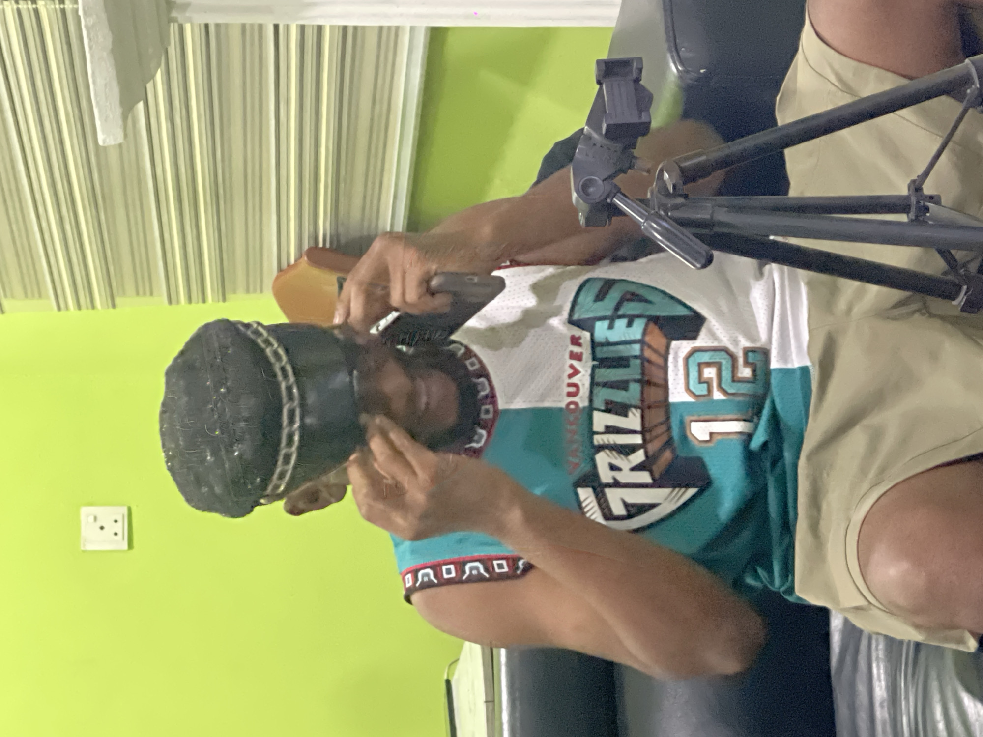 A man is wearing a black brimless hat and a Vancouver Grizzlies #12 jersey. He is holding a black object to his face, which appears to be a phone or recording device, while seated in what looks like a wheelchair. A black camera stand or microphone stand is positioned in front of him. The background consists of a light green wall with an electrical outlet, and vertical blinds. It is not possible to confirm the location in the city of Abuja, Nigeria.