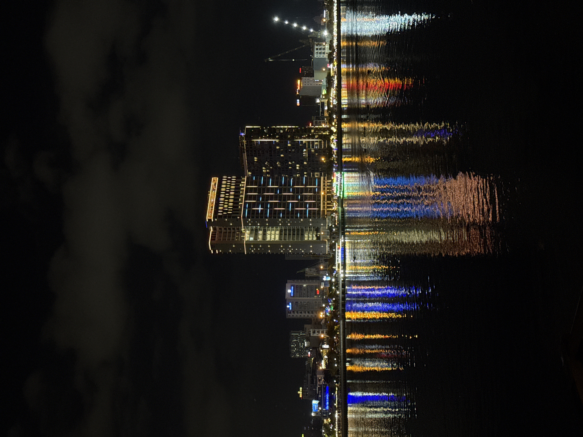 This is a night-time, long-exposure photograph of the Han River waterfront in Da Nang, Vietnam. The central focus is on a cluster of illuminated high-rise buildings, one of which is significantly taller and wider than the others, featuring a grid pattern of lights and a glowing yellow top. To its right, a slightly shorter building is also lit. The cityscape extends to the left and right with smaller buildings and what appears to be a construction crane with lights in the distance. The water in the foreground reflects the vibrant colors of the buildings and lights, creating a shimmering, abstract pattern of blue, yellow, and red streaks. The sky is dark and cloudy, with some faint illumination from the city. A boat with green lights is visible on the water, contributing to the reflections. The overall impression is one of a modern, active urban waterfront at night.